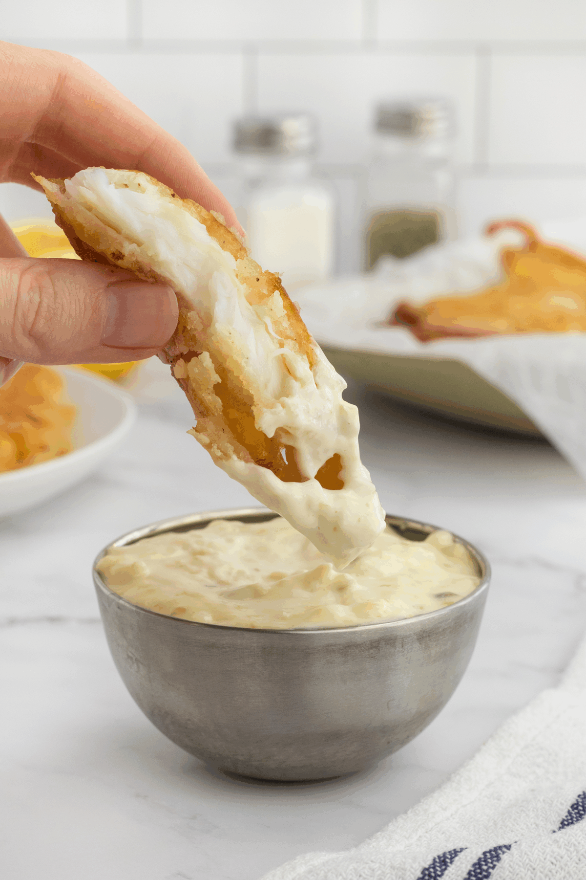 A hand dips a piece of battered, flaky fish into a bowl of creamy tartar sauce. In the background are salt and pepper shakers, a plate of more battered fish, and a white tiled wall.