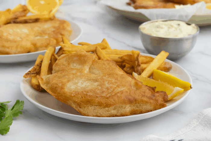 A plate with crispy battered fish, golden fries, and lemon wedges. A bowl of tartar sauce and another plate with fish and fries are in the background.