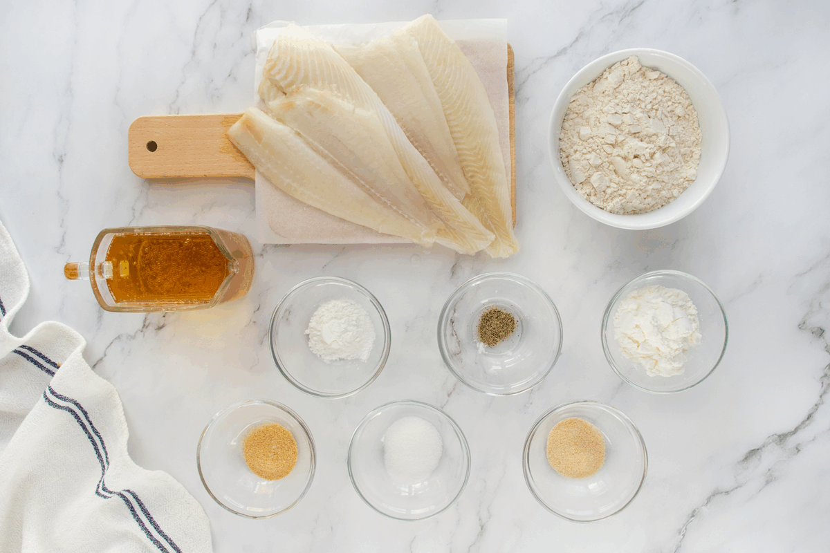 Overhead view of raw fish fillets on a small cutting board with bowls of flour, seasonings, cornstarch, sugar, and a bottle of honey on a white marble surface, next to a striped kitchen towel.