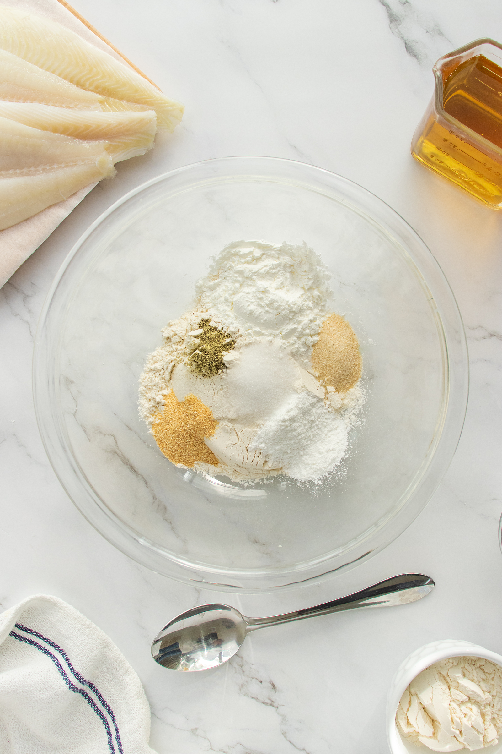 A glass bowl with flour, spices, and seasonings sits on a marble surface. Nearby are raw fish fillets, a jar of oil, a spoon, a folded towel, and a small bowl of flour.