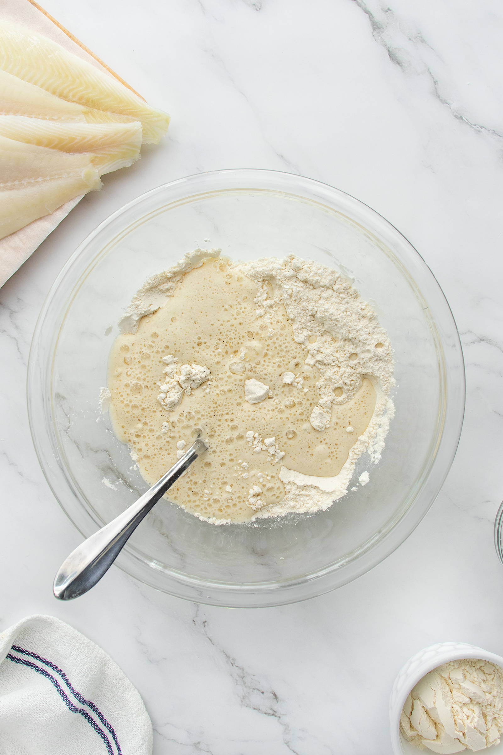 A glass bowl filled with flour and liquid mixture, partially stirred with a metal spoon, sits on a marble countertop. Striped towels and fish fillets are nearby.