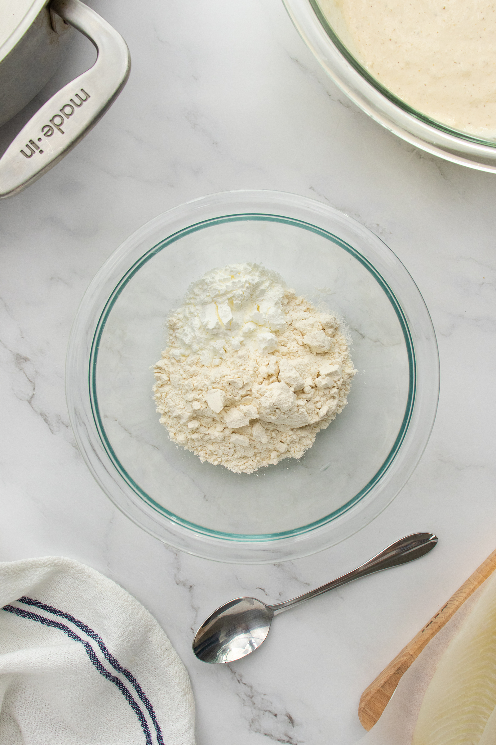 A clear glass bowl with flour and baking powder sits on a white marble countertop. Nearby are a metal spoon, a stand mixer, a striped kitchen towel, and a bowl with batter.