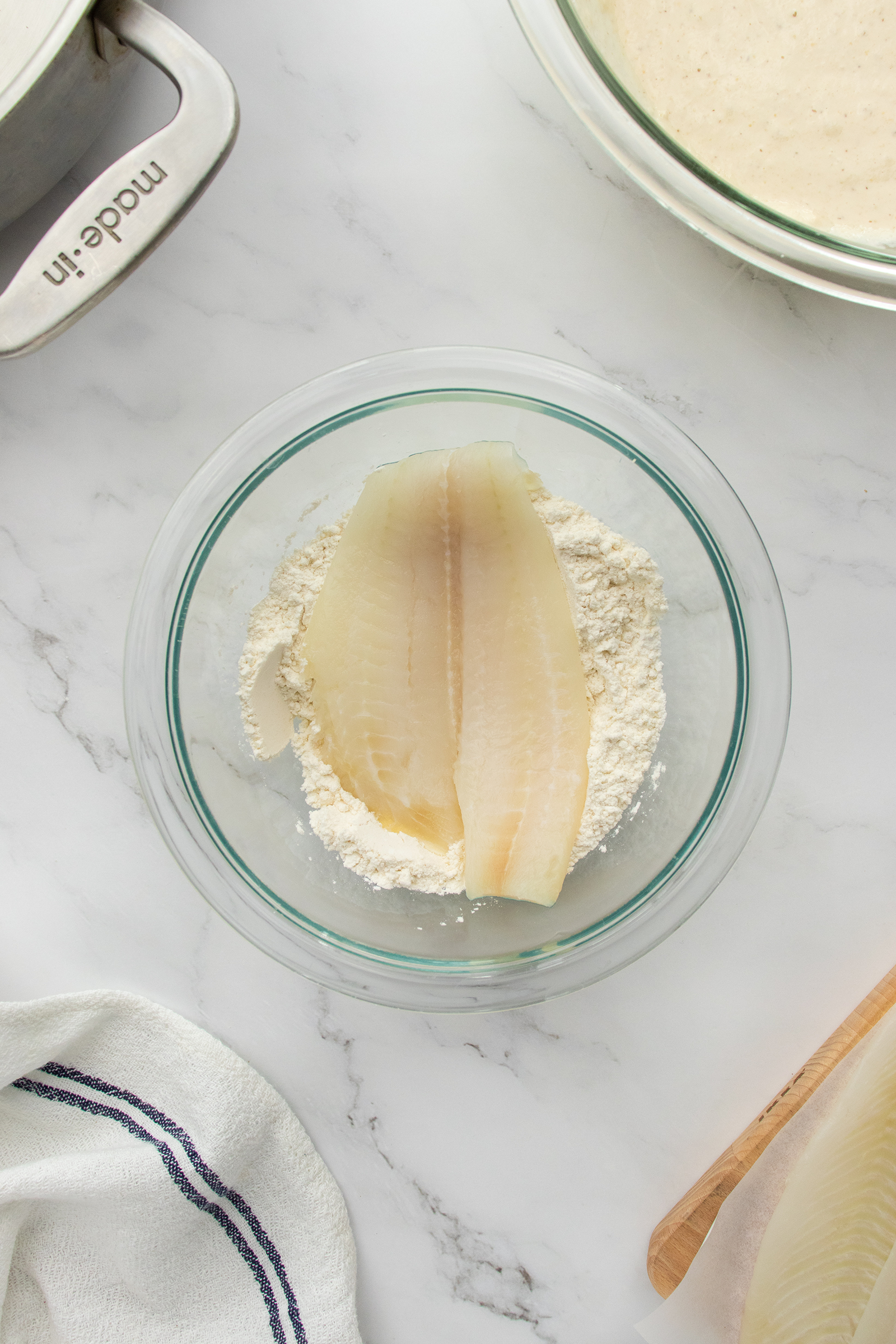 Two raw fish fillets sit in a glass bowl with flour on a white marble countertop. Nearby are a white towel with a navy stripe, a pan, and a bowl containing batter.