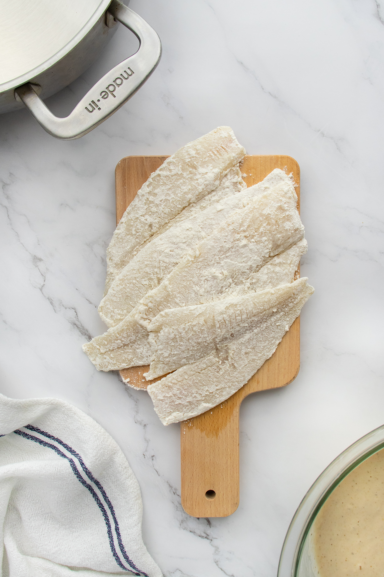 Three raw fish fillets coated in flour are arranged on a small wooden cutting board. A metal pot, a white towel, and part of a glass bowl are visible around the board on a white marble surface.