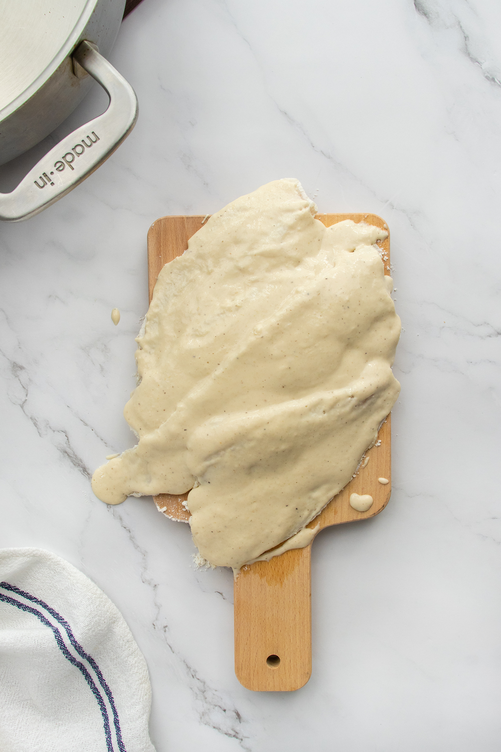 Three raw fish fillets coated in batter are arranged on a small wooden cutting board. A metal pot, a white towel, and part of a glass bowl are visible around the board on a white marble surface.