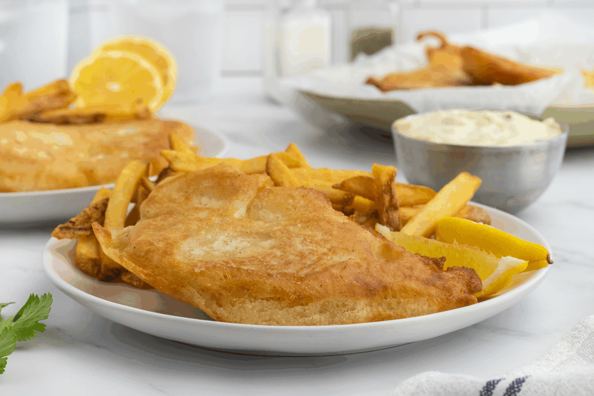 A plate of crispy battered fish fillet with golden French fries is served with a lemon wedge. In the background, there’s a bowl of tartar sauce, another plate of fish and chips, and a cut lemon.