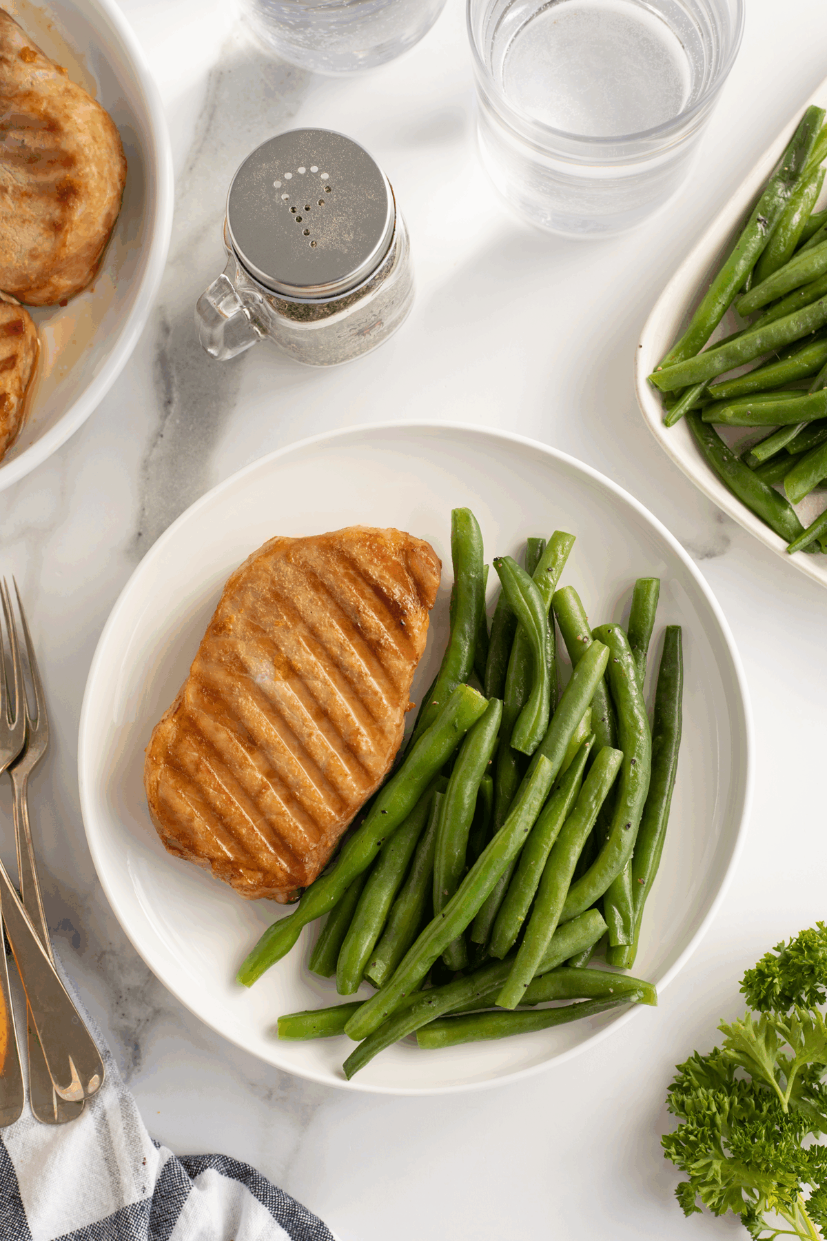 A plate with a grilled pork chop and a serving of steamed green beans. Nearby are a dish of salt, two glasses of water, a serving plate of green beans, silverware, and a sprig of parsley on a white surface.
