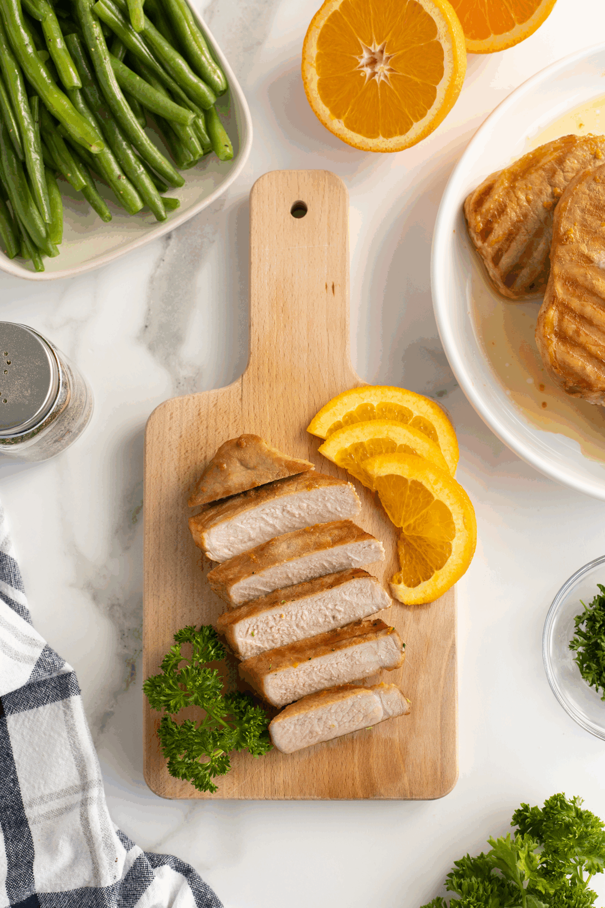 Sliced cooked pork chop on a wooden cutting board with parsley and orange slices, surrounded by green beans, orange halves, pepper, and a white napkin on a marble countertop.