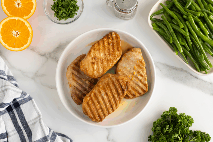 Three grilled pork chops in a white bowl on a marble surface, surrounded by a dish of green beans, fresh parsley, a halved orange, salt shaker, and a striped kitchen towel.