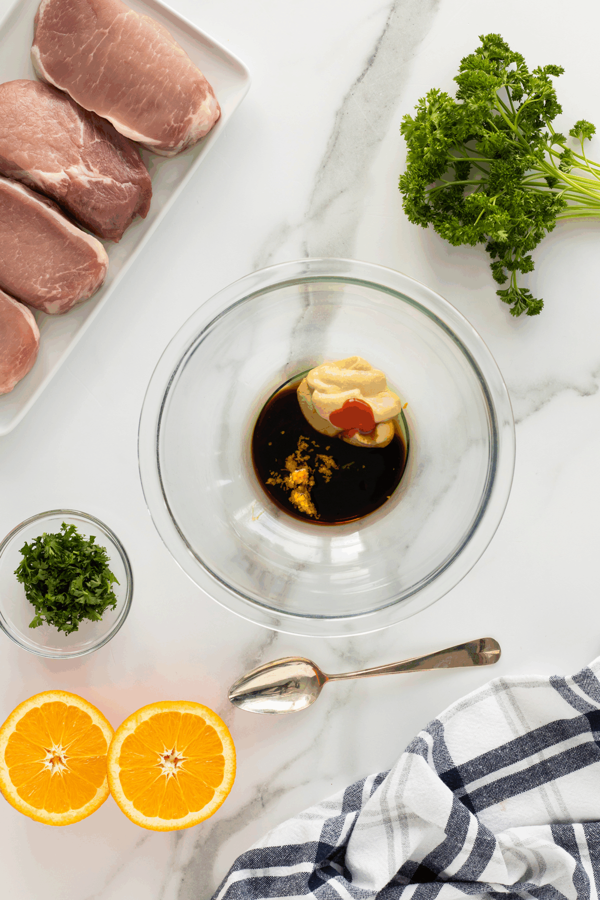 A glass bowl with soy sauce, mustard, and a dab of red sauce sits on a marble counter near raw pork chops, fresh parsley, orange halves, chopped herbs, a spoon, and a striped cloth.