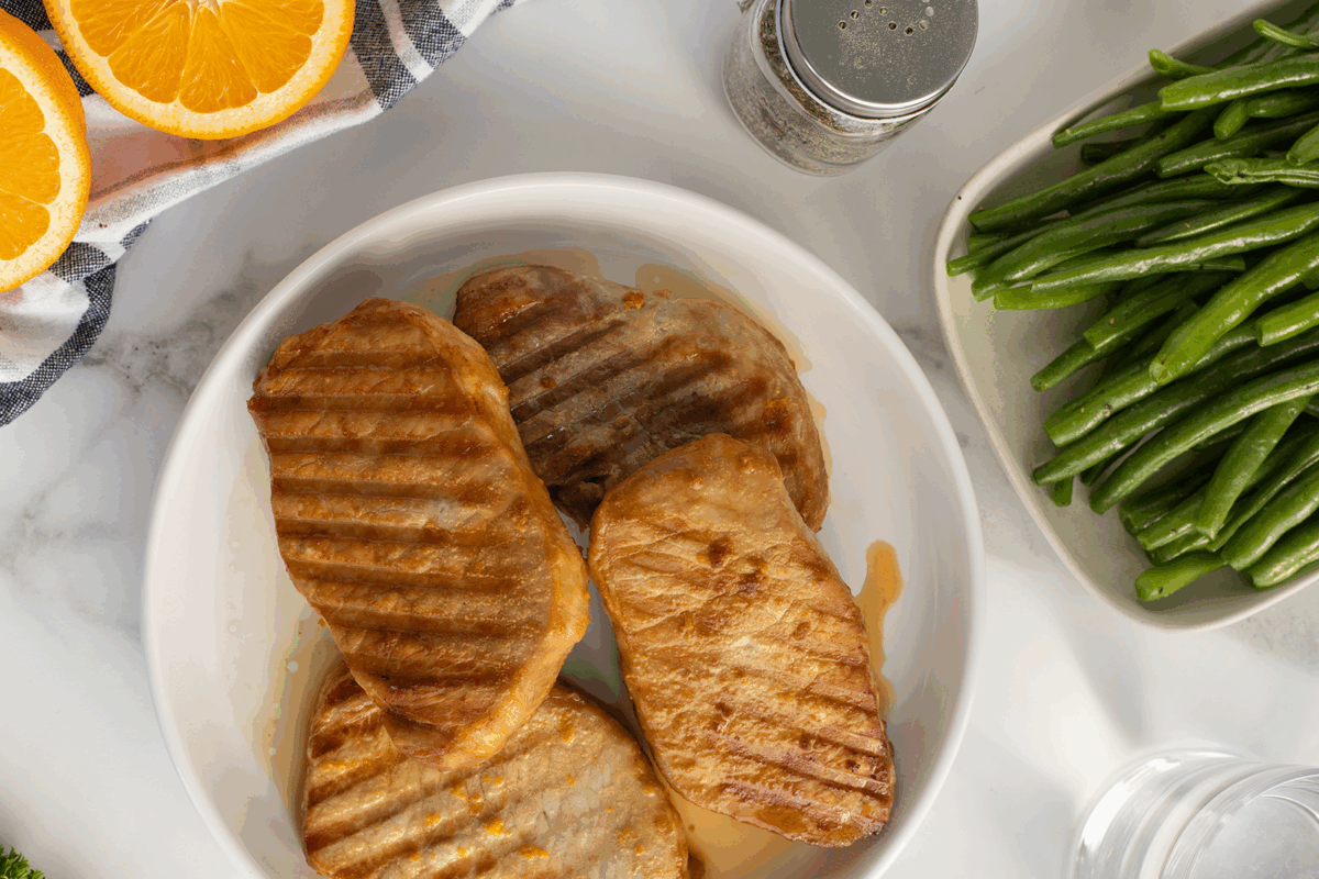 A white plate with three grilled pork chops, next to a dish of green beans, orange slices, a pepper shaker, and part of a glass of water on a marble surface.