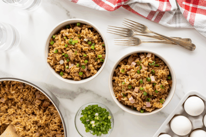 Two bowls of fried rice with chopped green onions and ham on a marble surface, surrounded by a skillet of rice, a bowl of green onions, four forks, eggs, two glasses of water, and a red-and-white checkered towel.