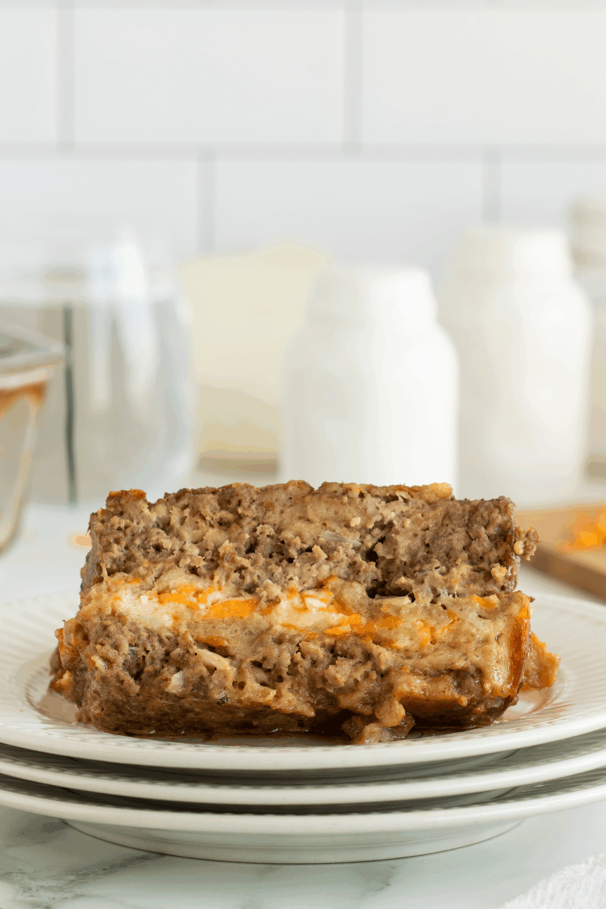 A close-up of a slice of meatloaf with a layer of melted cheese in the center, served on a stack of white plates with blurred kitchen jars in the background.