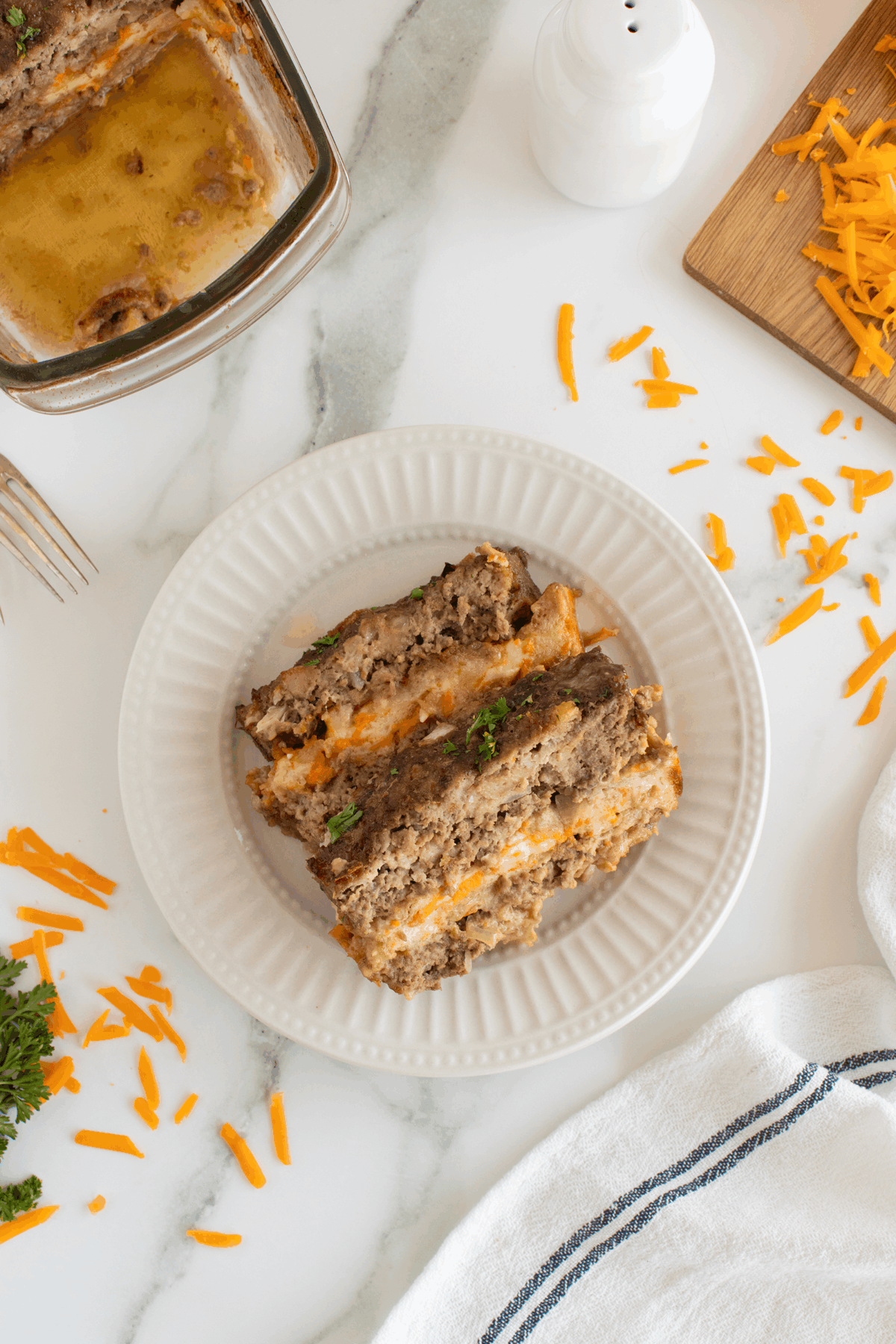 Two slices of meatloaf with melted cheese are served on a white plate. Shredded cheddar cheese and a cutting board are nearby, with a glass baking dish containing more meatloaf in the background.