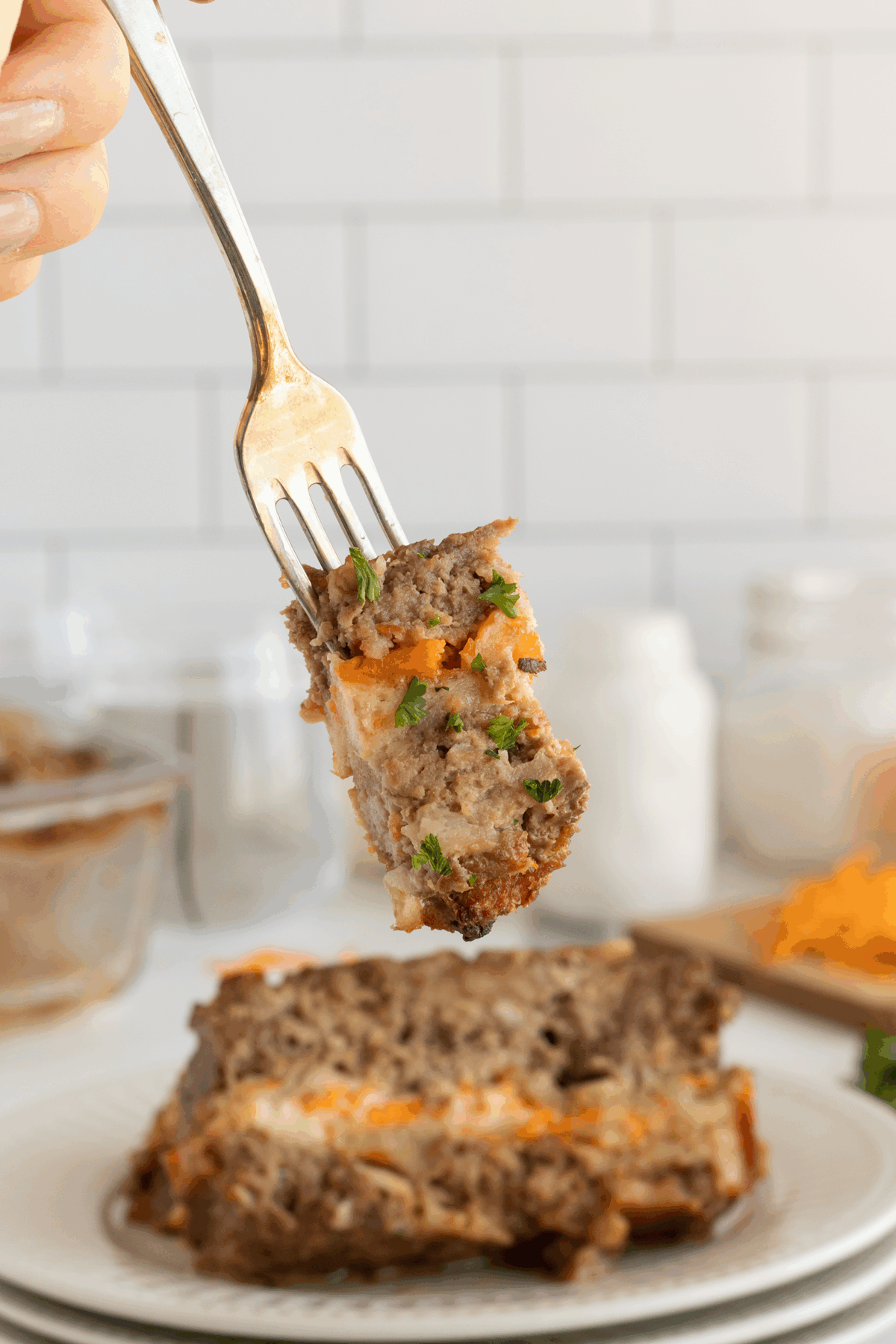 A fork holds a bite of layered meatloaf with cheese and herbs above a plate with a large slice of meatloaf. The background includes blurred jars and shredded cheese on a white counter.
