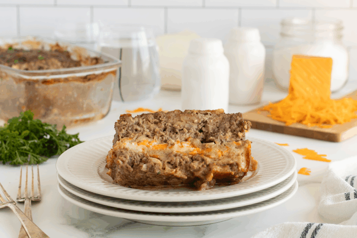 Two slices of cheesy meatloaf are stacked on a white plate. In the background, there’s a baking dish with more meatloaf, shredded cheese on a cutting board, parsley, and salt and pepper shakers on a kitchen counter.