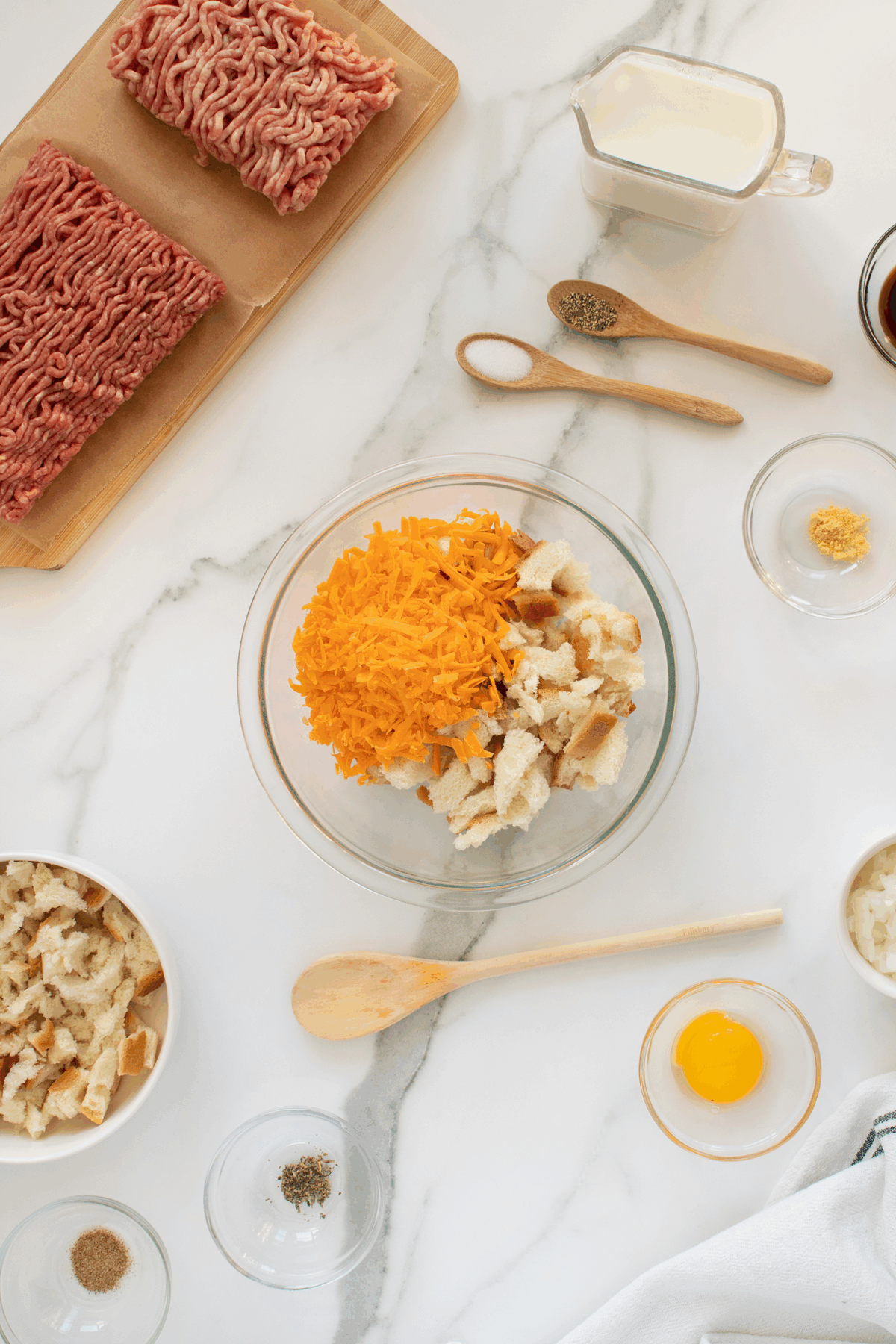 Top-down view of meatloaf ingredients on a marble counter: ground beef, shredded cheese, bread pieces, egg, milk, chopped onion, and seasonings, with utensils and small bowls arranged neatly around a mixing bowl.