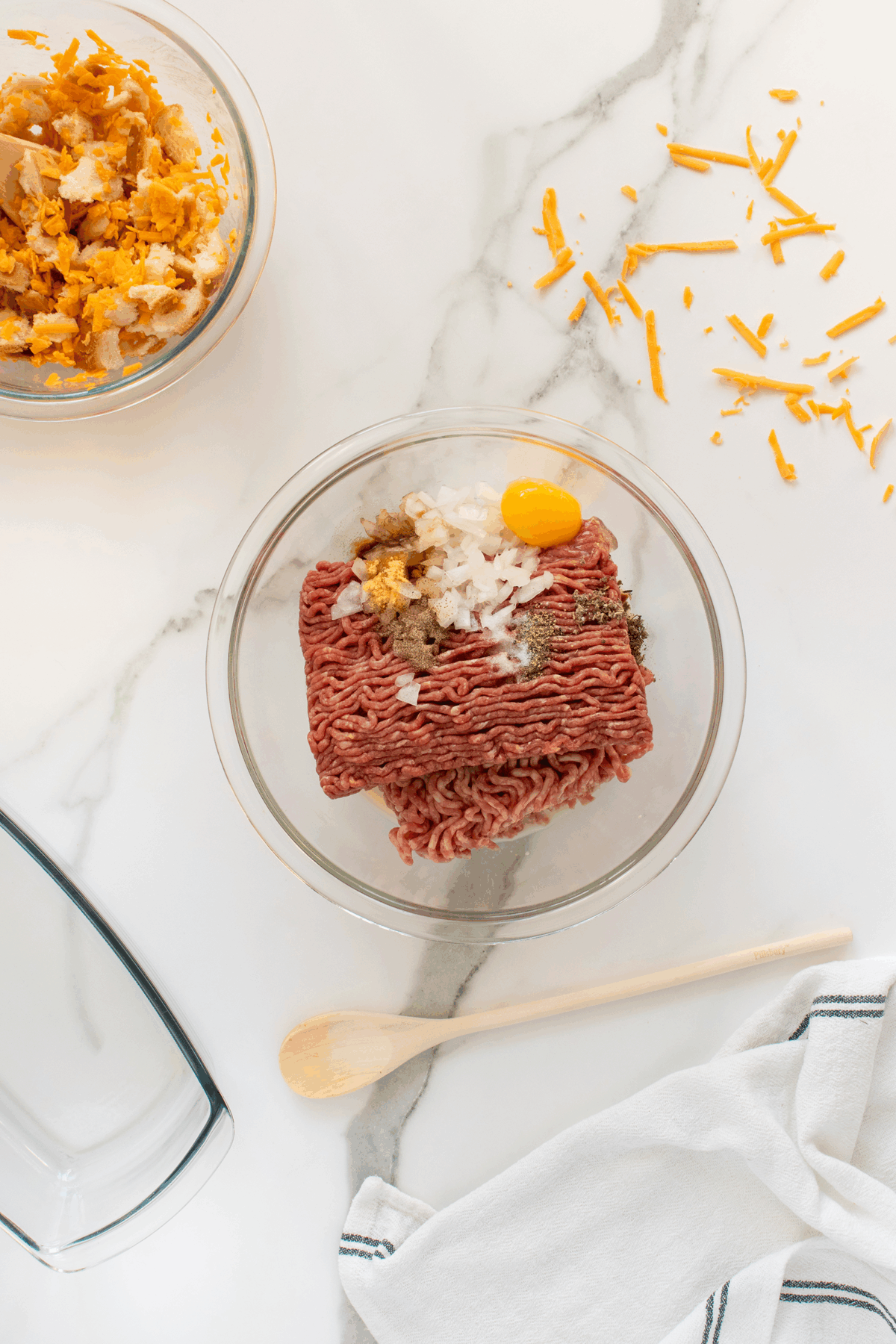 A glass bowl with ground beef, a cracked egg, chopped onions, and seasonings sits on a marble countertop. Nearby are a bowl of crushed crackers, shredded cheddar cheese, a wooden spoon, and a white towel.