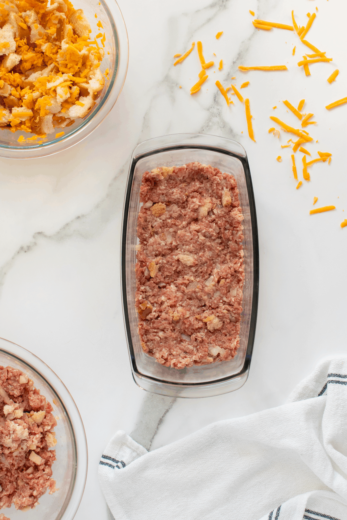 A glass loaf pan filled with uncooked meatloaf mixture sits on a marble surface. Surrounding the pan are bowls with breadcrumbs and shredded cheese, and some cheese is scattered on the counter. A white towel is nearby.