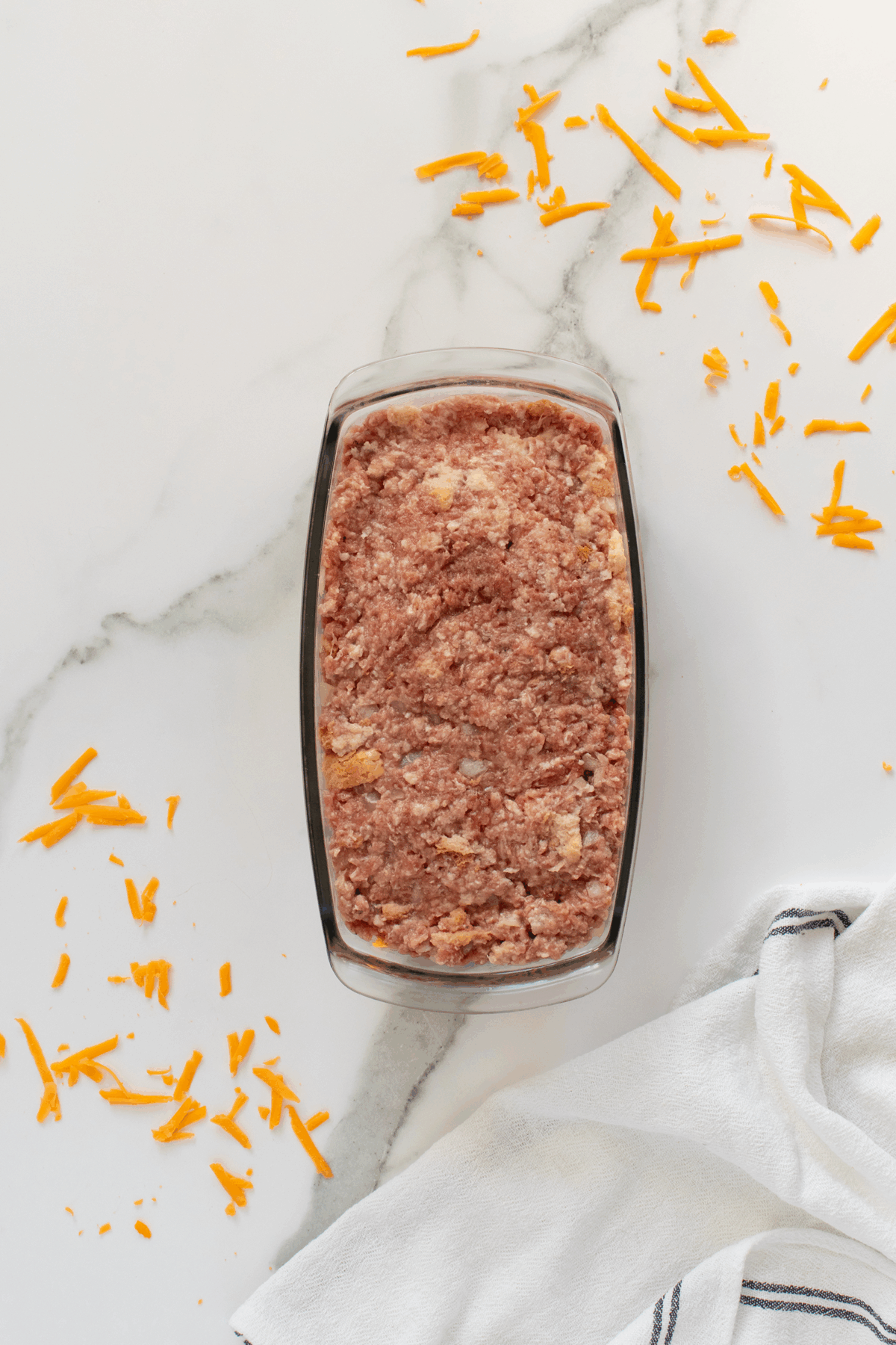 A glass loaf pan filled with uncooked meatloaf mixture sits on a marble surface, surrounded by shredded cheddar cheese and a white towel with dark stripes.