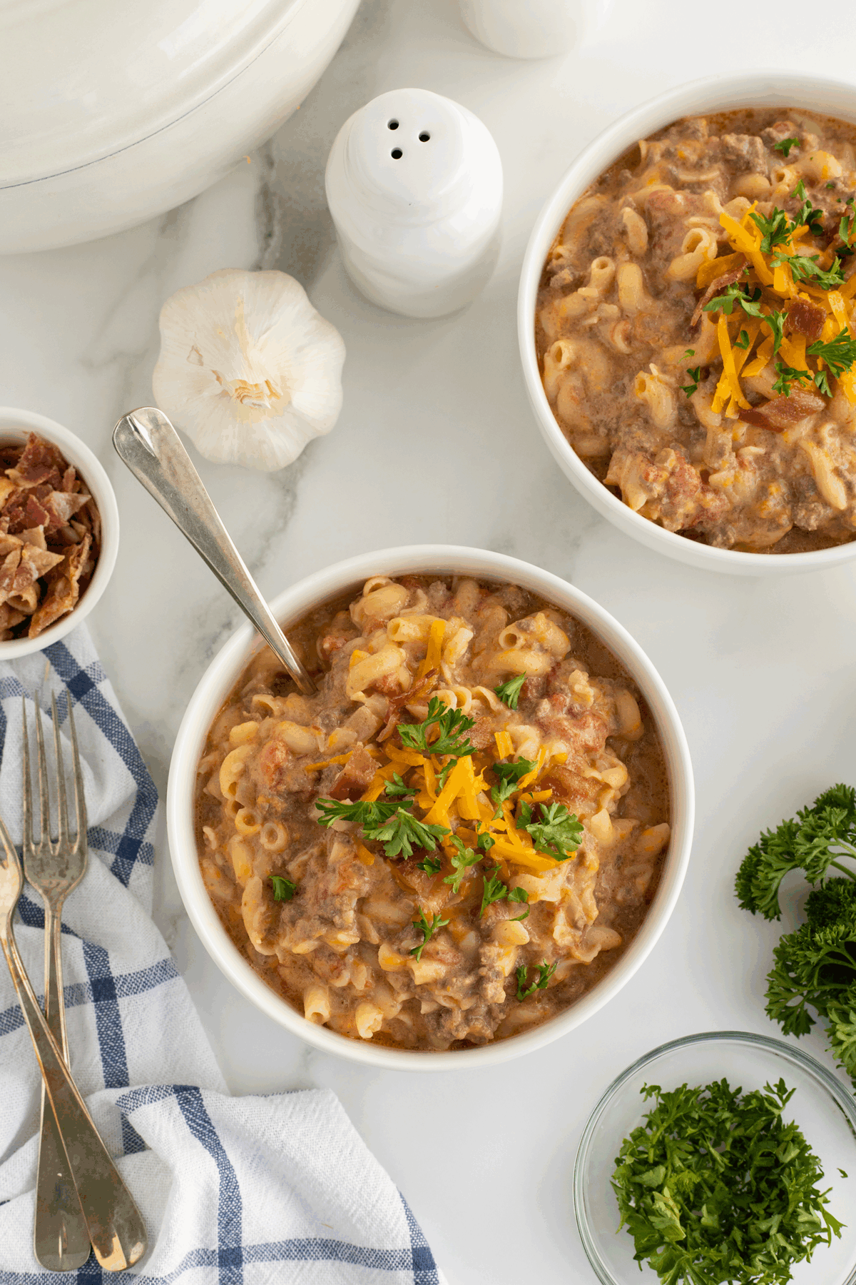Two bowls of cheesy macaroni and ground beef topped with shredded cheese and parsley, set on a white surface with a garlic bulb, salt shaker, bacon bits, forks, a striped towel, and fresh parsley nearby.
