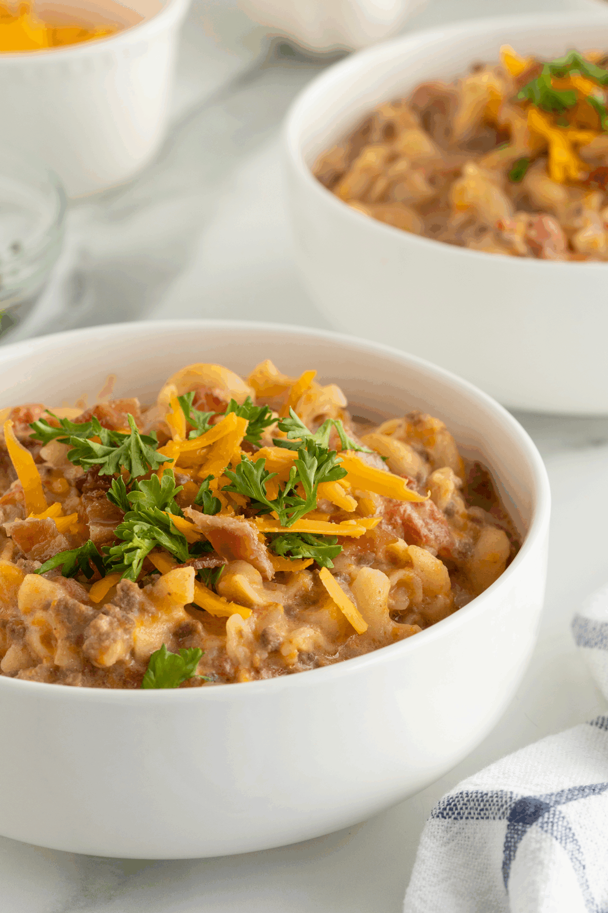 A white bowl filled with creamy beef and pasta, topped with shredded cheddar cheese and chopped parsley, sits on a white surface with another similar bowl in the background.