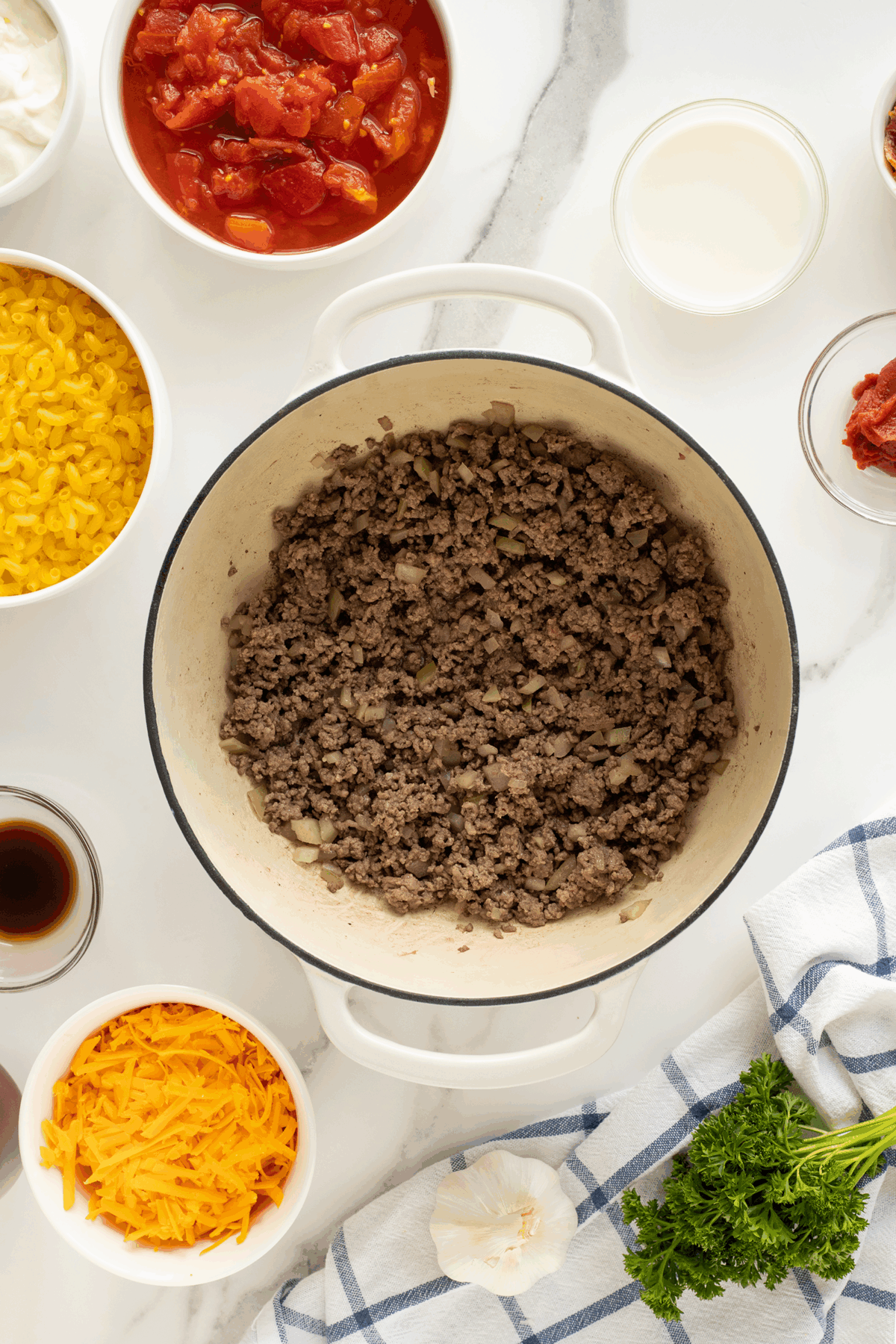 A white pot filled with cooked ground beef and onions sits on a marble countertop, surrounded by bowls of shredded cheese, macaroni, tomatoes, milk, tomato paste, Worcestershire sauce, and fresh parsley with a kitchen towel.