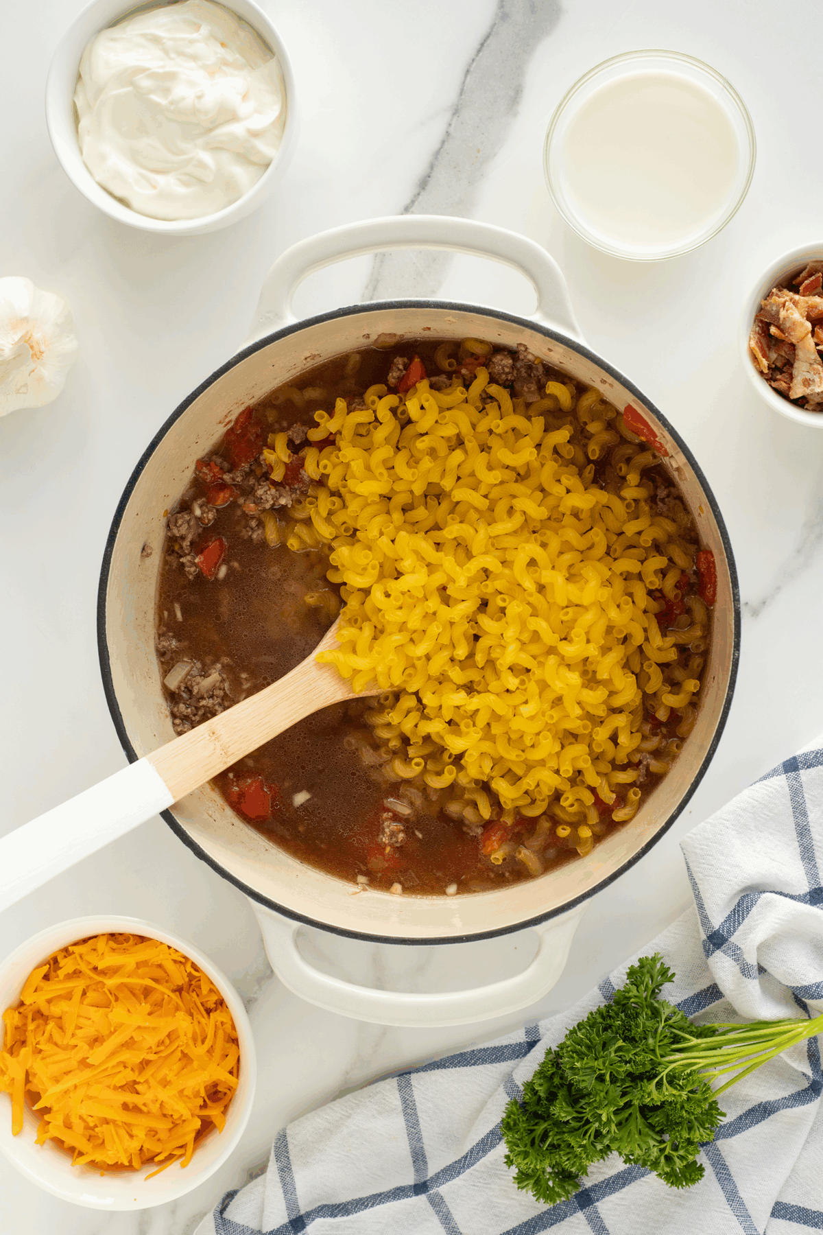 A pot of uncooked macaroni, ground meat, and tomatoes is being stirred with a wooden spoon. Surrounding the pot are bowls of shredded cheese, sour cream, milk, bacon, garlic, and fresh parsley on a white countertop.