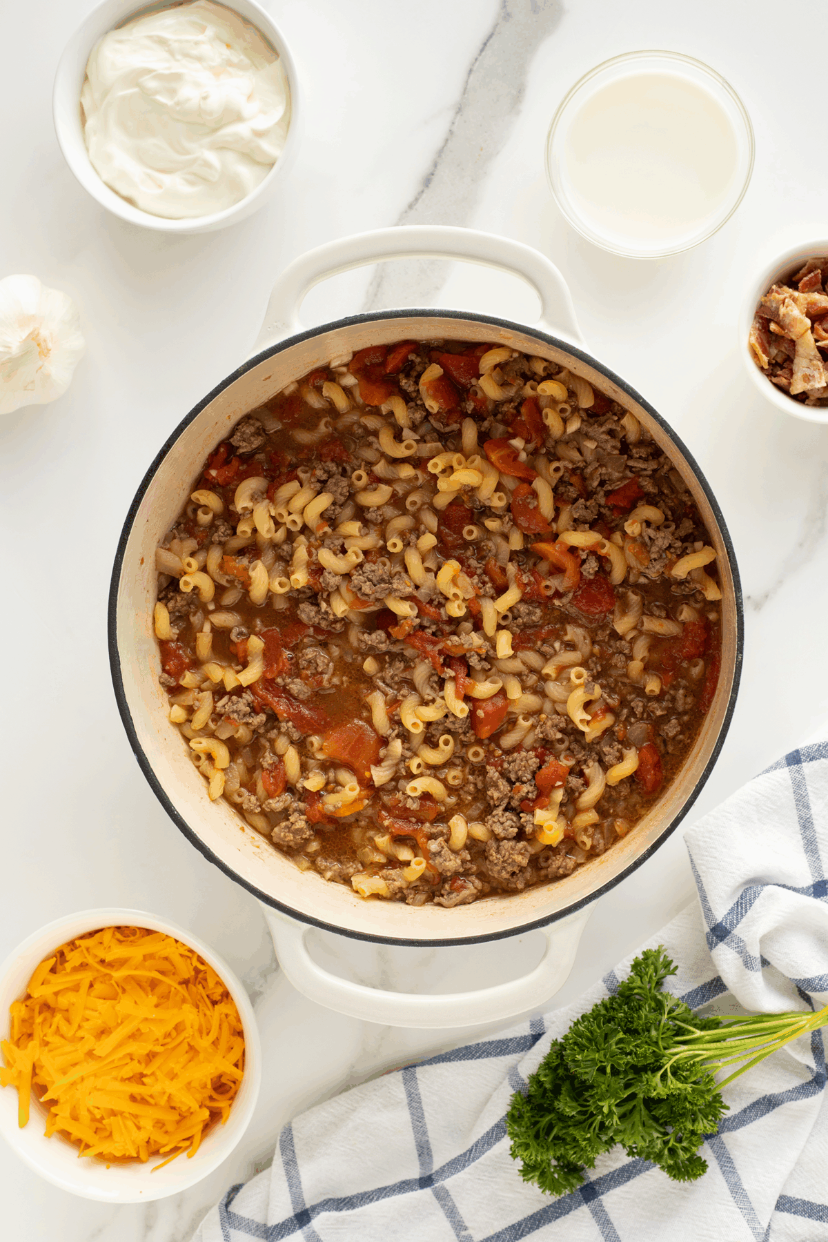 A pot of cooked macaroni, ground beef, and tomatoes sits on a white counter, surrounded by bowls of shredded cheddar cheese, sour cream, milk, parsley, bacon, a garlic bulb, and a blue-and-white striped towel.