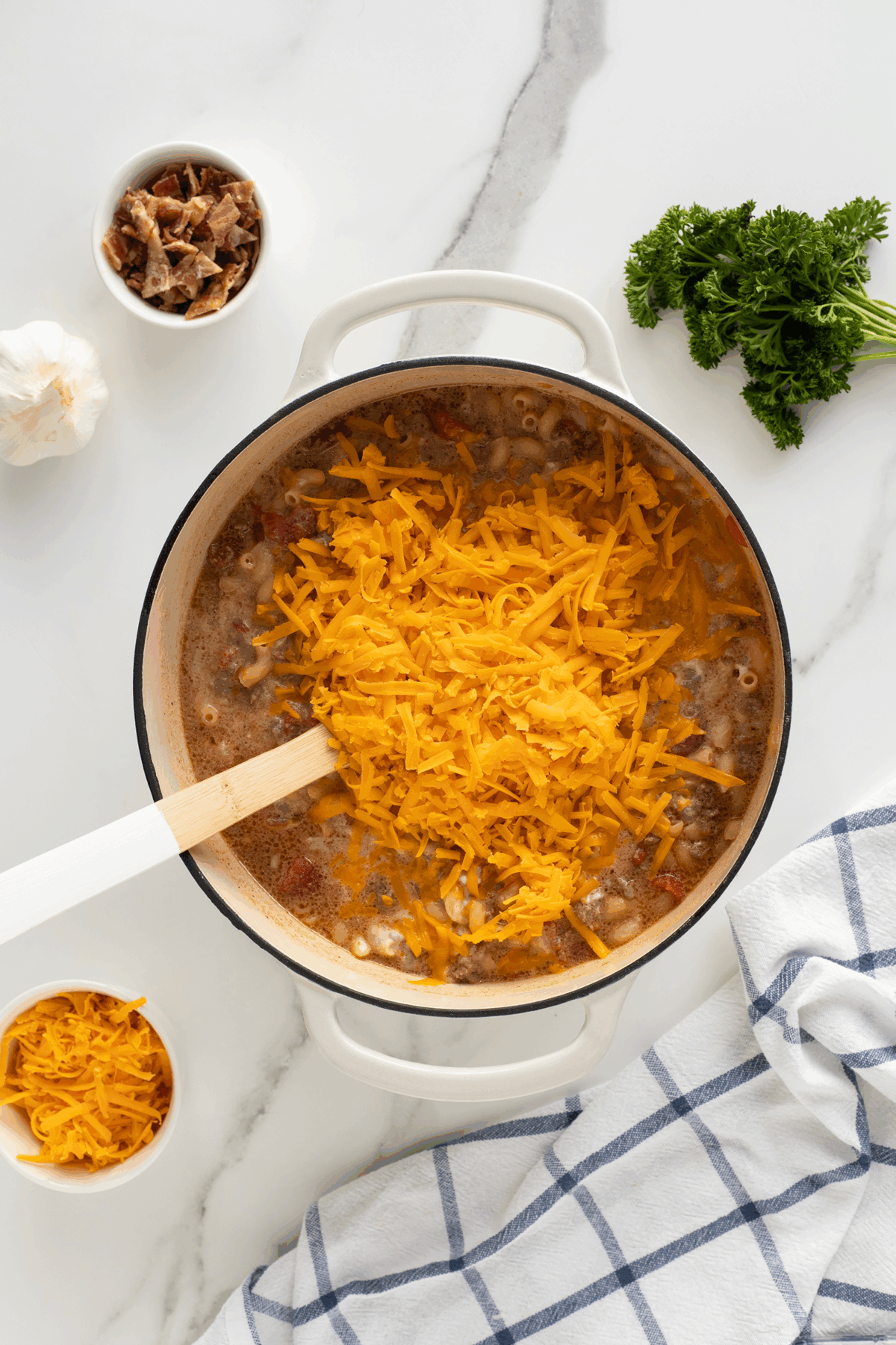 A white pot filled with soup and topped with shredded cheddar cheese sits on a marble surface. Nearby are bowls of bacon bits and shredded cheese, a bunch of parsley, a garlic bulb, and a striped kitchen towel.