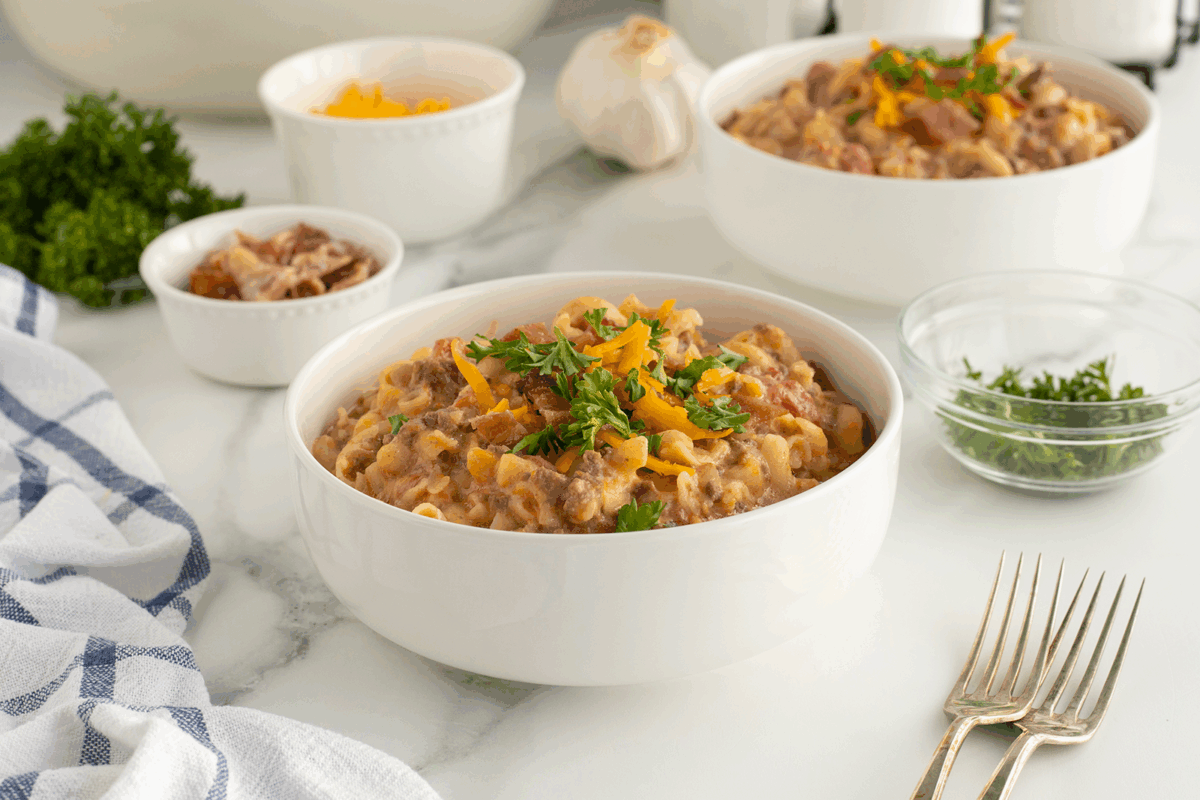 A white bowl filled with cheesy pasta and ground beef, topped with fresh parsley and shredded cheese, sits on a marble counter with a towel, two forks, and small bowls of garnishes in the background.