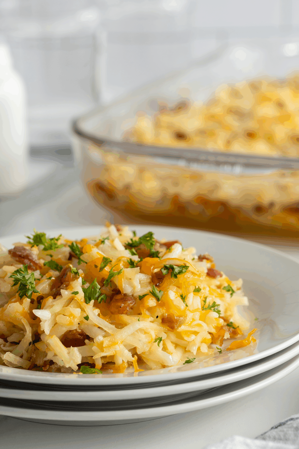 A close-up of a plate of cheesy hash brown casserole topped with chopped parsley, with a glass baking dish filled with more casserole in the background.