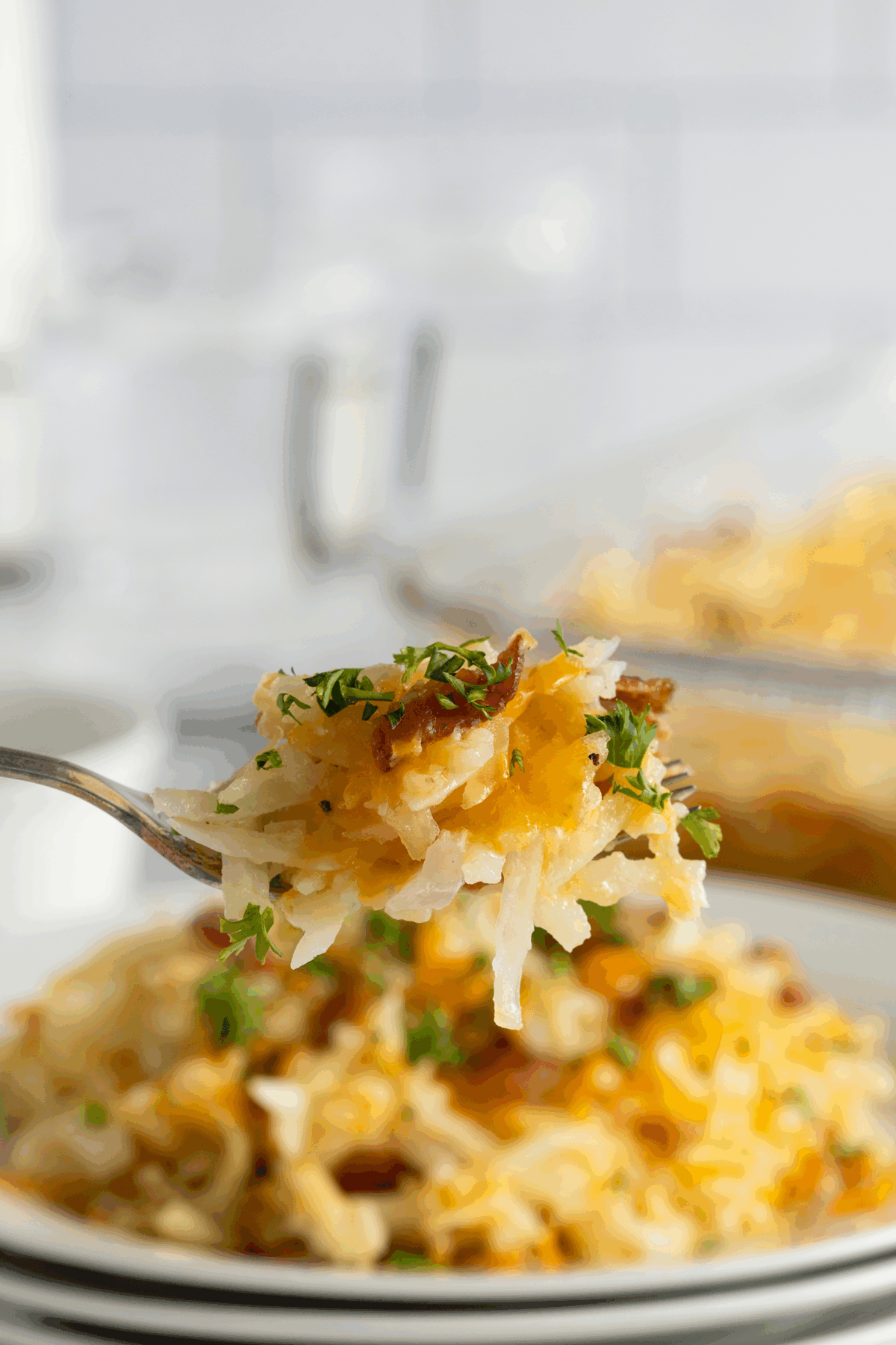 A close-up of a fork holding a bite of cheesy hash brown casserole garnished with fresh herbs, with a plate and baking dish of the casserole blurred in the background.