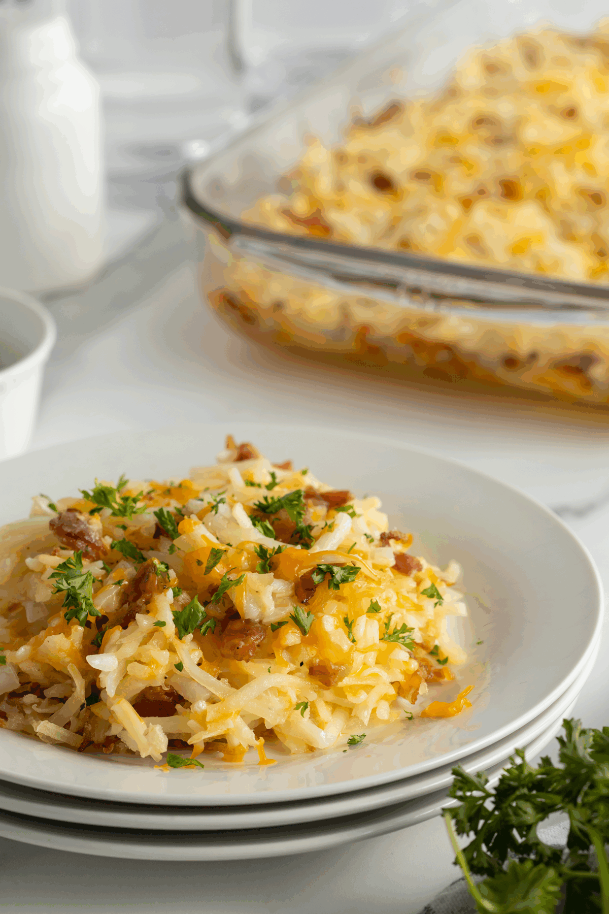 A serving of cheesy hash brown casserole garnished with parsley is on a white plate, with a larger casserole dish of the same dish in the background.