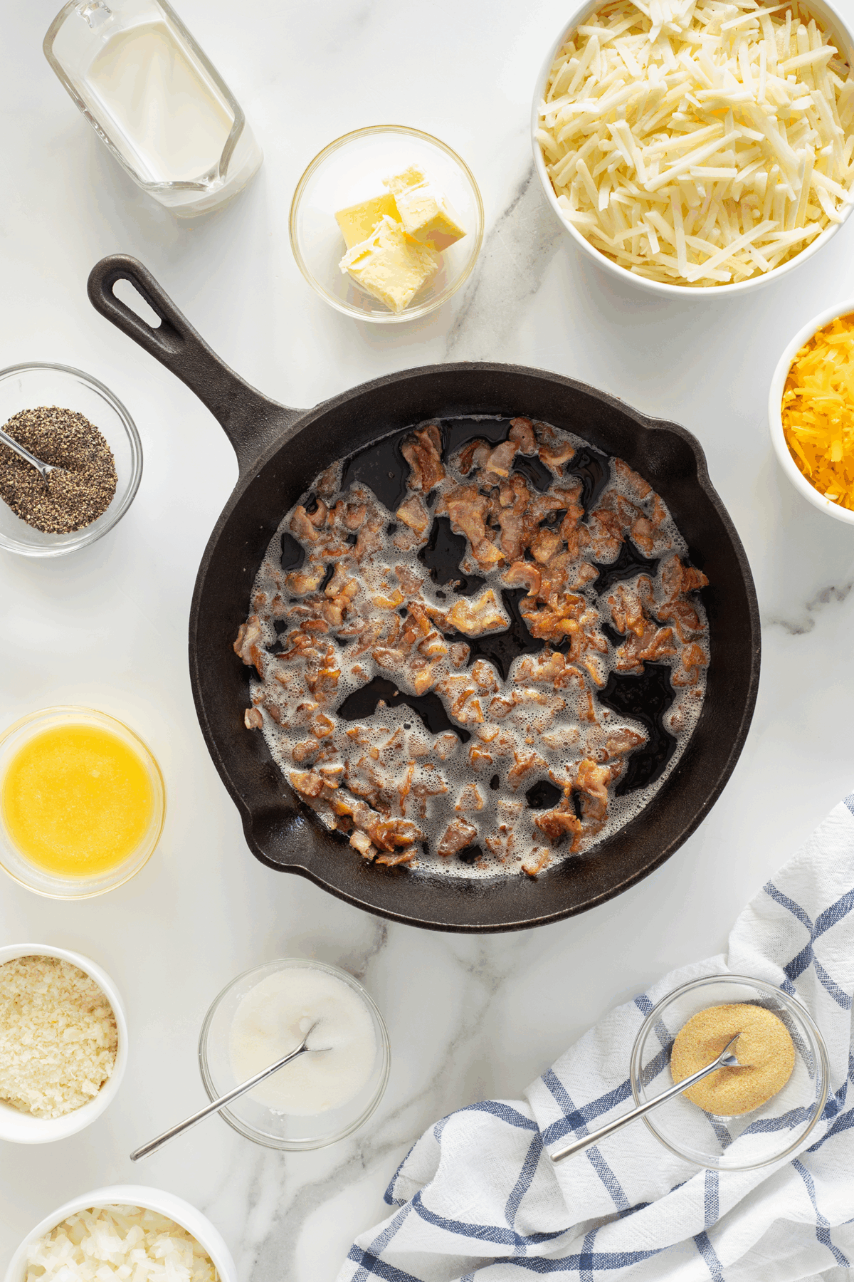 A cast iron skillet with cooked bacon pieces sits on a marble surface, surrounded by bowls of shredded potatoes, shredded cheese, cream, butter, melted butter, sour cream, seasonings, and a blue-striped kitchen towel.