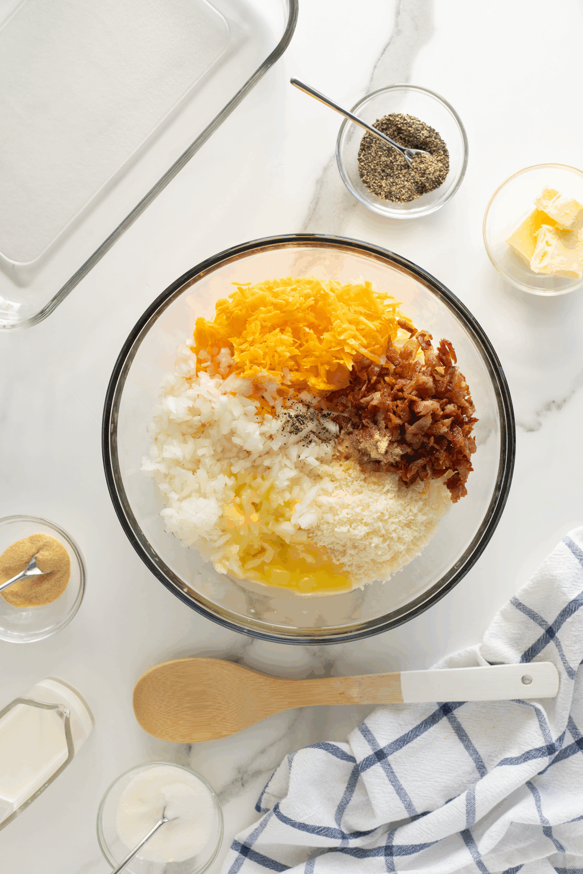 A glass bowl with shredded cheese, chopped onions, bacon, egg, and seasonings sits on a marble counter surrounded by small bowls of spices, a butter dish, a wooden spoon, a baking dish, and a blue checked towel.