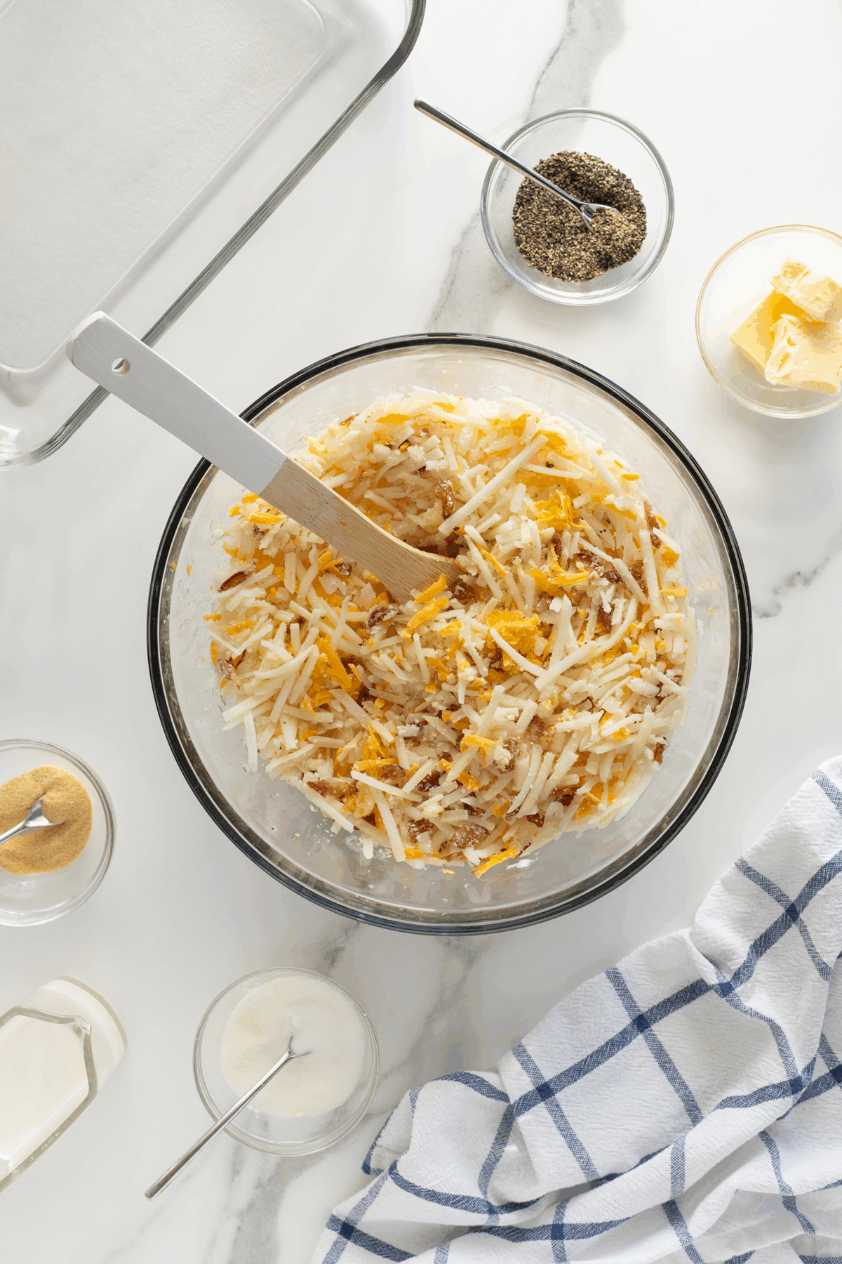 A glass bowl filled with shredded potatoes and cheese being mixed, surrounded by small bowls of spices, butter, milk, and a white dish towel on a white countertop.