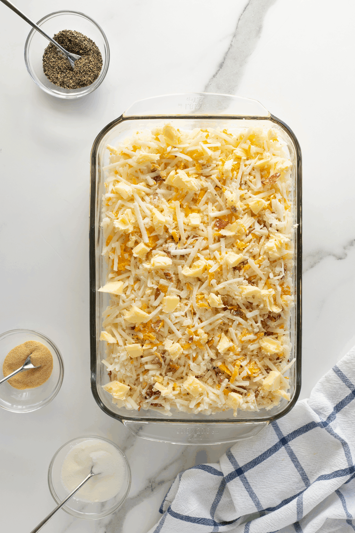 A glass baking dish filled with shredded potatoes and cheese sits on a marble counter. Nearby are bowls of pepper, garlic powder, and salt with spoons, and a blue-and-white checkered kitchen towel.