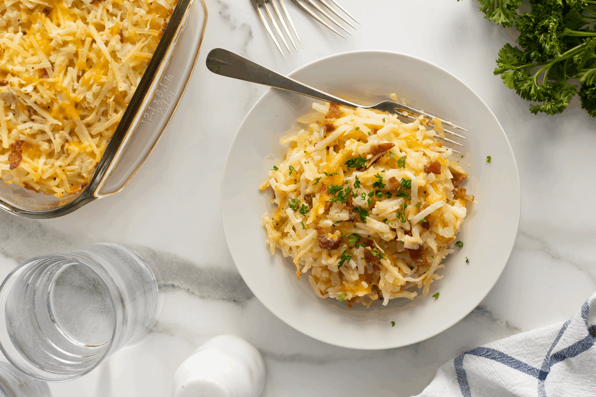 A plate of cheesy hash brown casserole garnished with parsley, with a fork on the plate. A baking dish with more casserole, water glasses, parsley, and a napkin are nearby on a white marble surface.