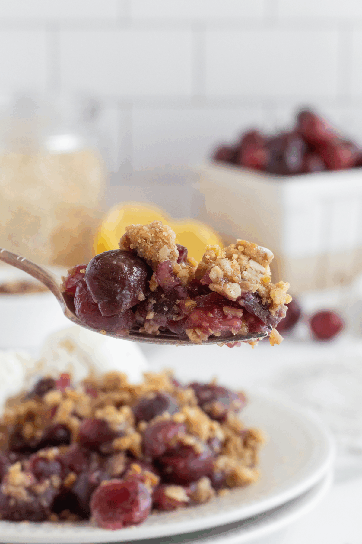A close-up of a spoon holding a serving of cherry crisp with oats, with a plate of cherry crisp, fresh cherries, and lemons in the blurred background.
