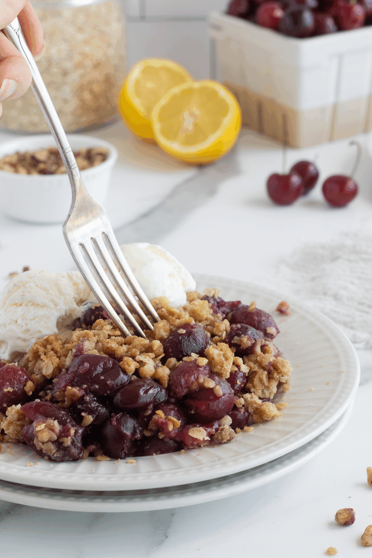 A fork cuts into a serving of cherry crumble with a crunchy topping, served with two scoops of vanilla ice cream on a white plate. Fresh cherries, lemon halves, and a container of oats are in the background.