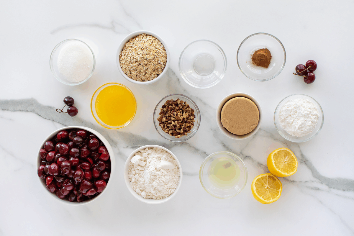Baking ingredients on a marble surface, including bowls of fresh cherries, oats, flour, sugar, nuts, melted butter, brown sugar, lemon halves, cinnamon, and clear liquids in small bowls.