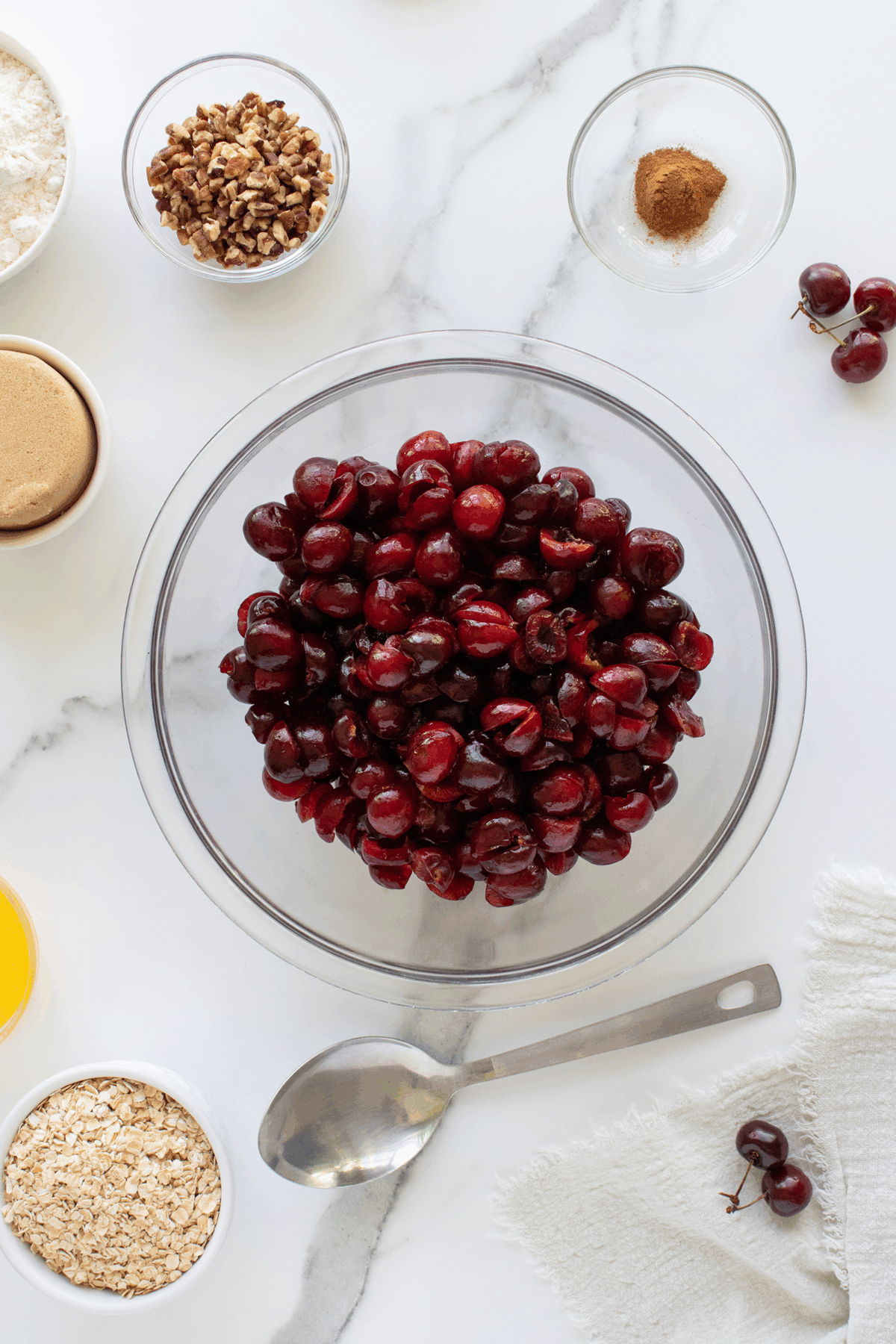 A glass bowl filled with pitted cherries sits on a white marble surface, surrounded by small bowls of brown sugar, flour, oats, chopped nuts, cinnamon, and a metal spoon.