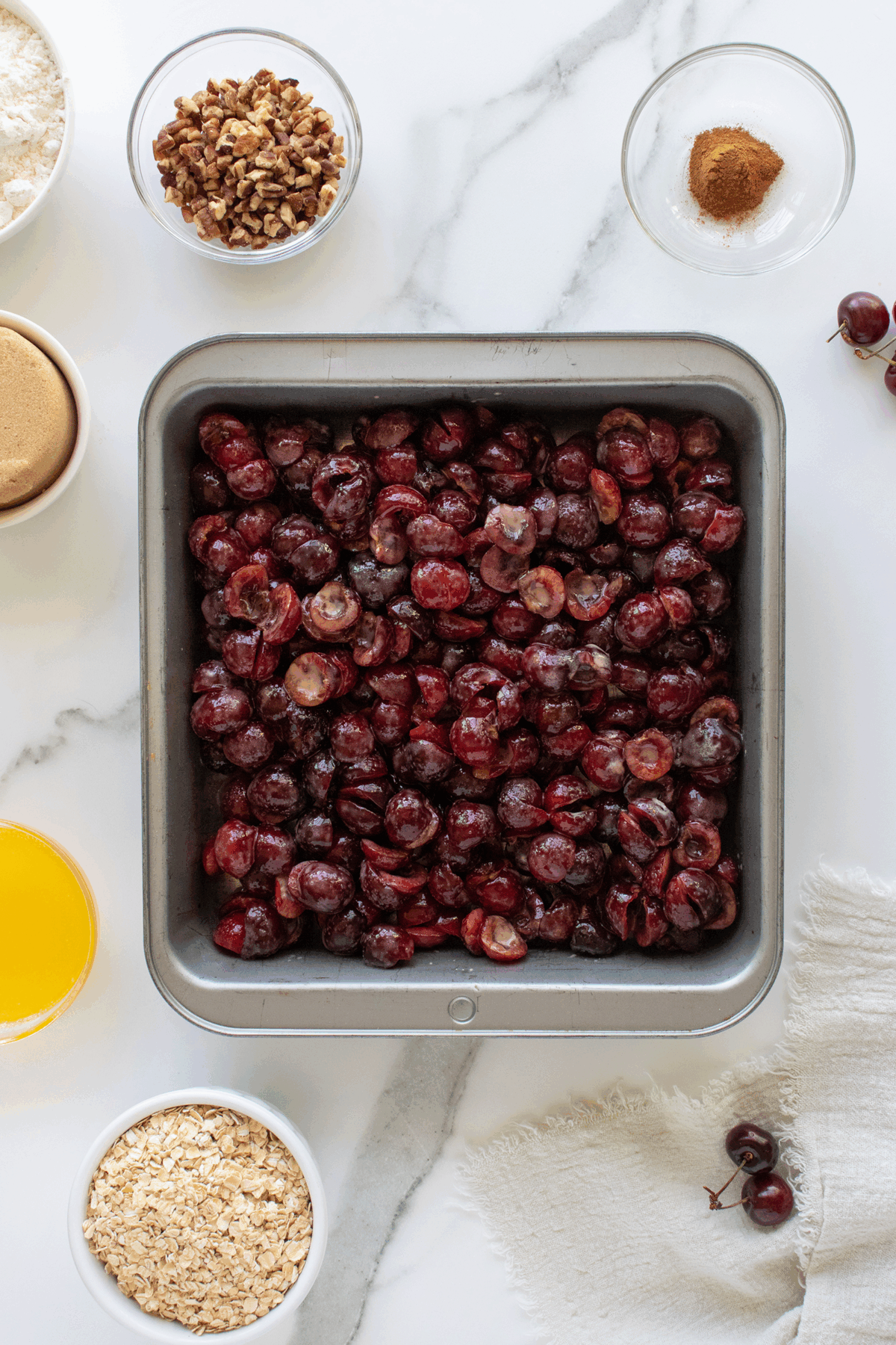 A square metal pan filled with halved cherries sits on a marble surface, surrounded by bowls containing chopped pecans, ground cinnamon, brown sugar, melted butter, oats, and flour.