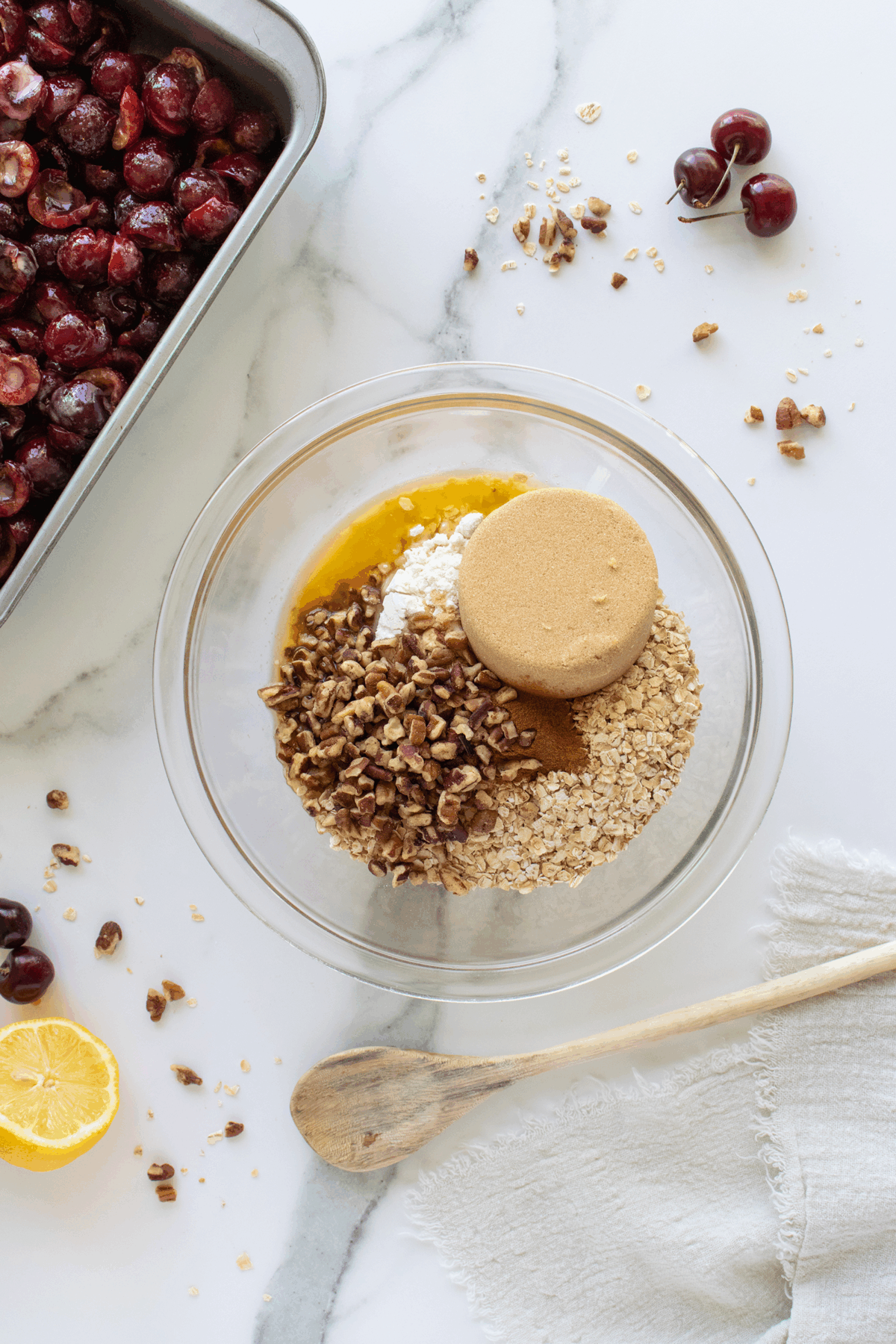 A glass bowl with oats, brown sugar, chopped pecans, flour, and cinnamon sits on a marble surface next to a wooden spoon. Nearby are a baking pan of halved cherries, a lemon half, and scattered pecans and cherries.
