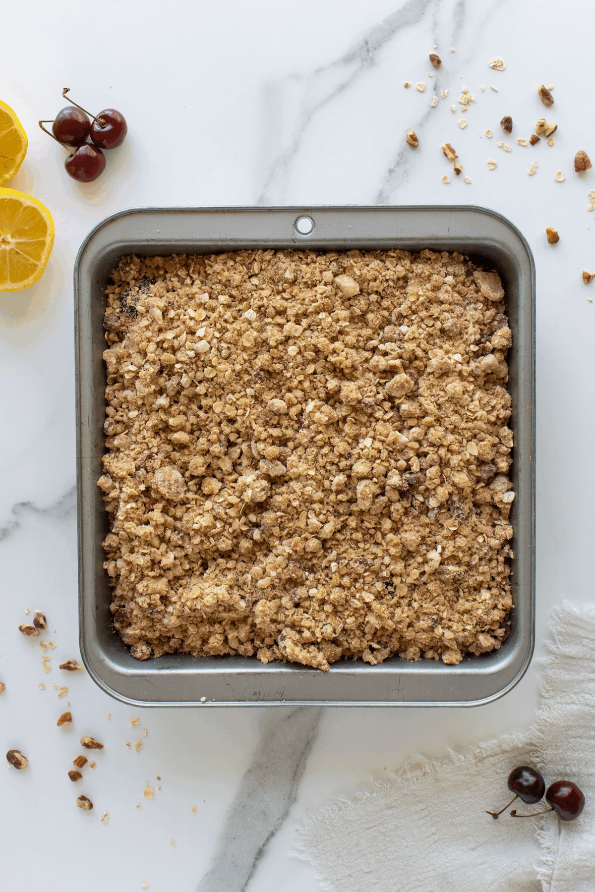 A square baking pan filled with golden brown fruit crumble sits on a marble surface, surrounded by lemon halves, cherries, oats, nuts, and a white cloth.