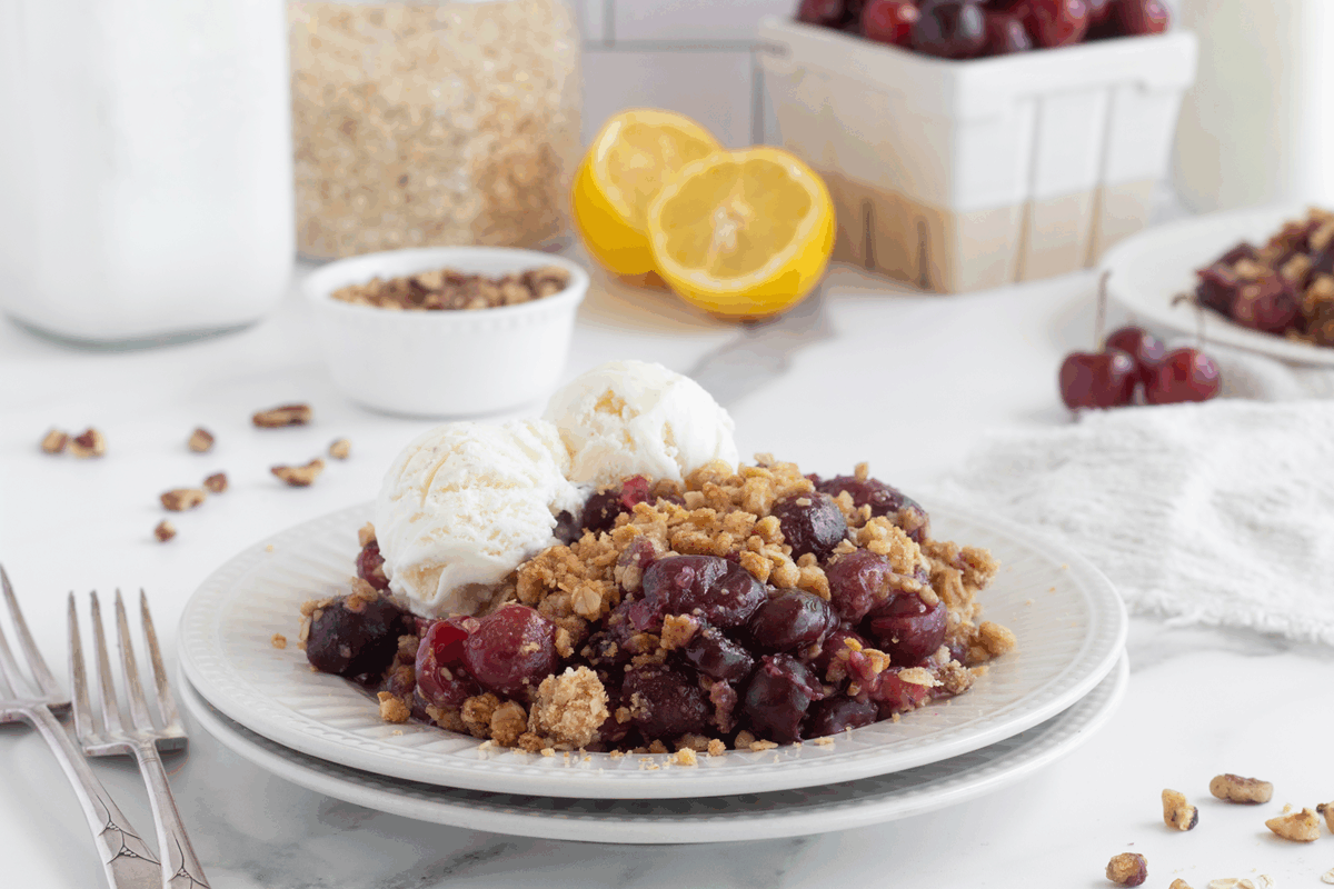 A plate of cherry crisp topped with two scoops of vanilla ice cream sits on a white table. Fresh cherries, lemon halves, and nuts are in the background, along with a fork and a white napkin.