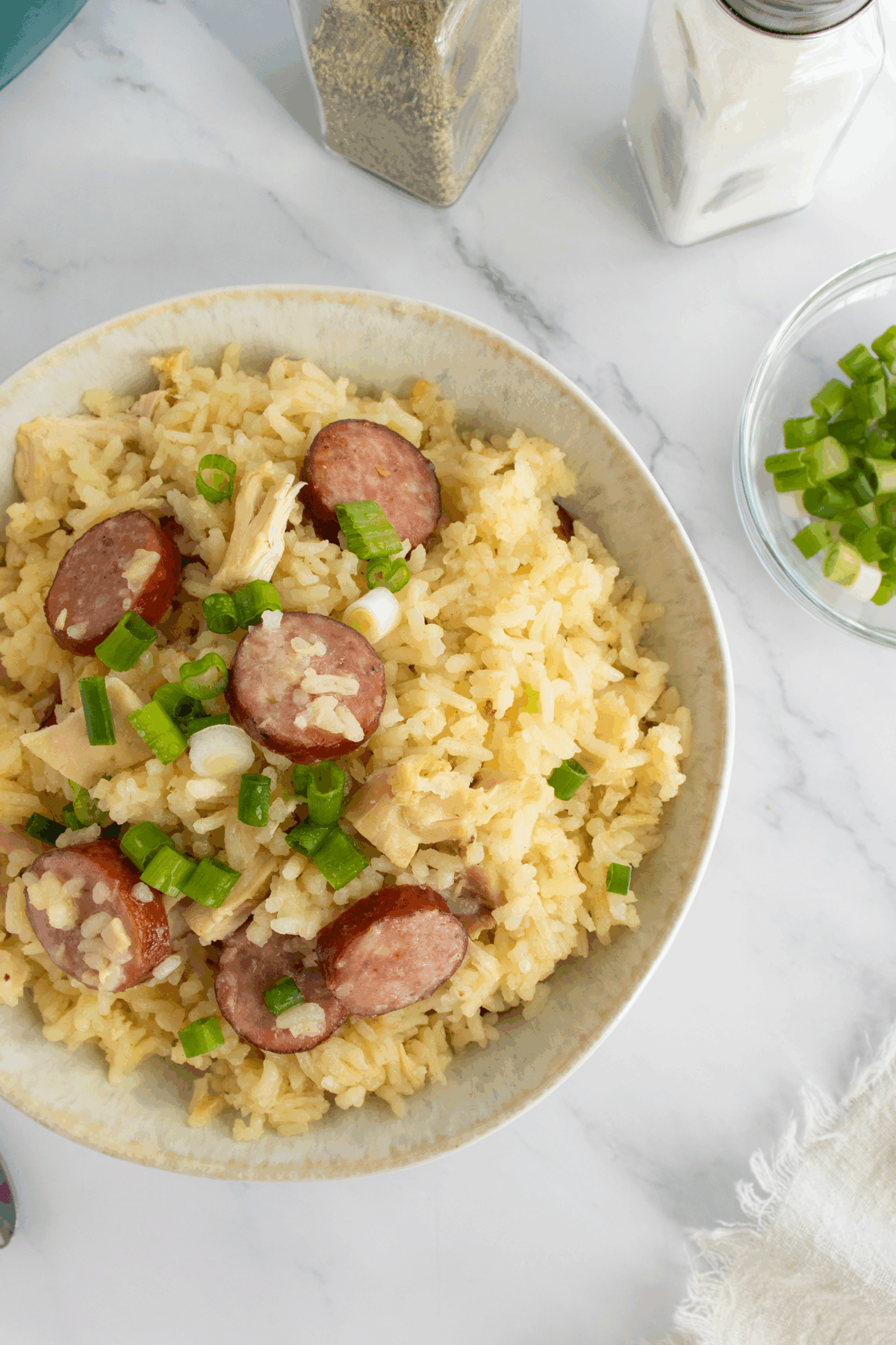 A bowl of rice mixed with sliced sausage, chicken pieces, and topped with chopped green onions, placed on a marble surface with a bowl of green onions and shakers of pepper and salt nearby.