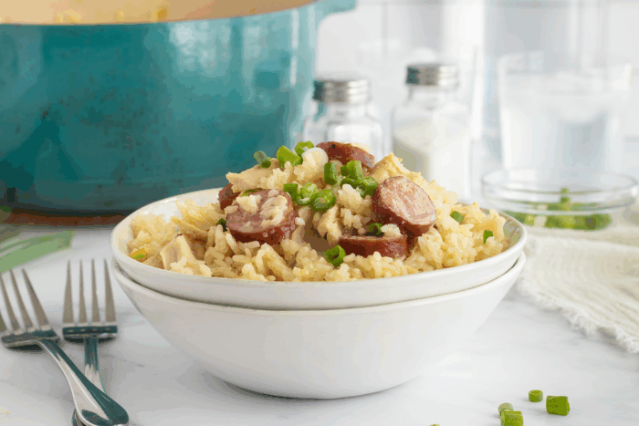 A white bowl filled with rice, sliced sausage, shredded chicken, and chopped green onions sits on a table beside forks and a blue pot, with salt and pepper shakers in the background.