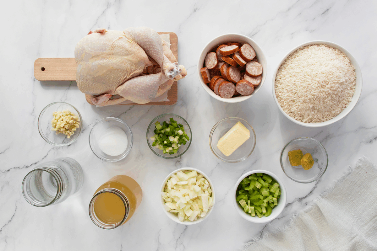 Raw whole chicken on a cutting board, sliced sausage, bowl of rice, chopped onions, celery, green peppers, minced garlic, butter, bouillon cubes, water, broth, and salt arranged on a white marble surface.
